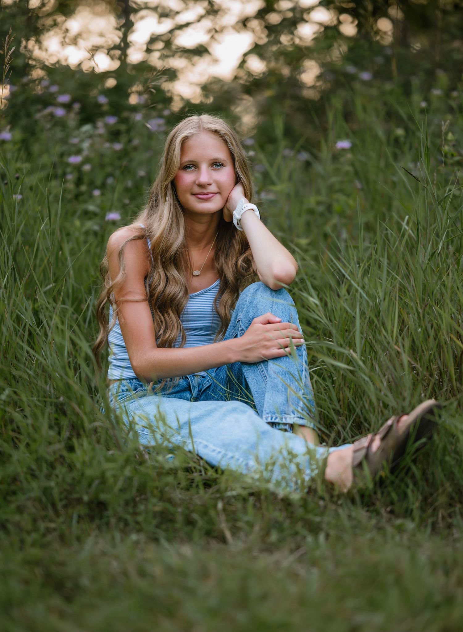 A person in a denim dress relaxes in tall summer grass during a golden hour outdoor portrait session.