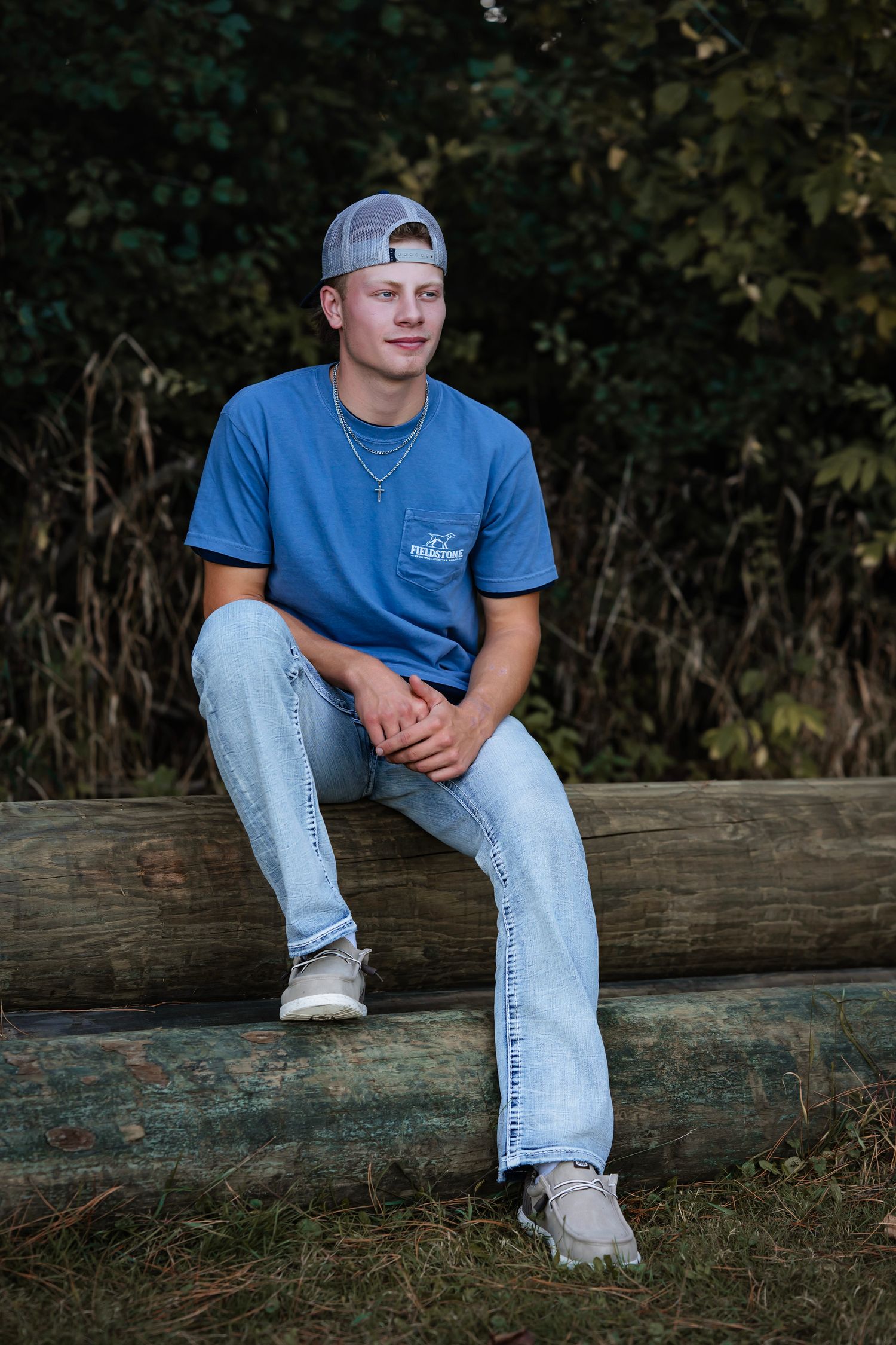 A casual portrait of someone wearing a blue t-shirt, baseball cap and jeans while sitting on a fallen log outdoors.