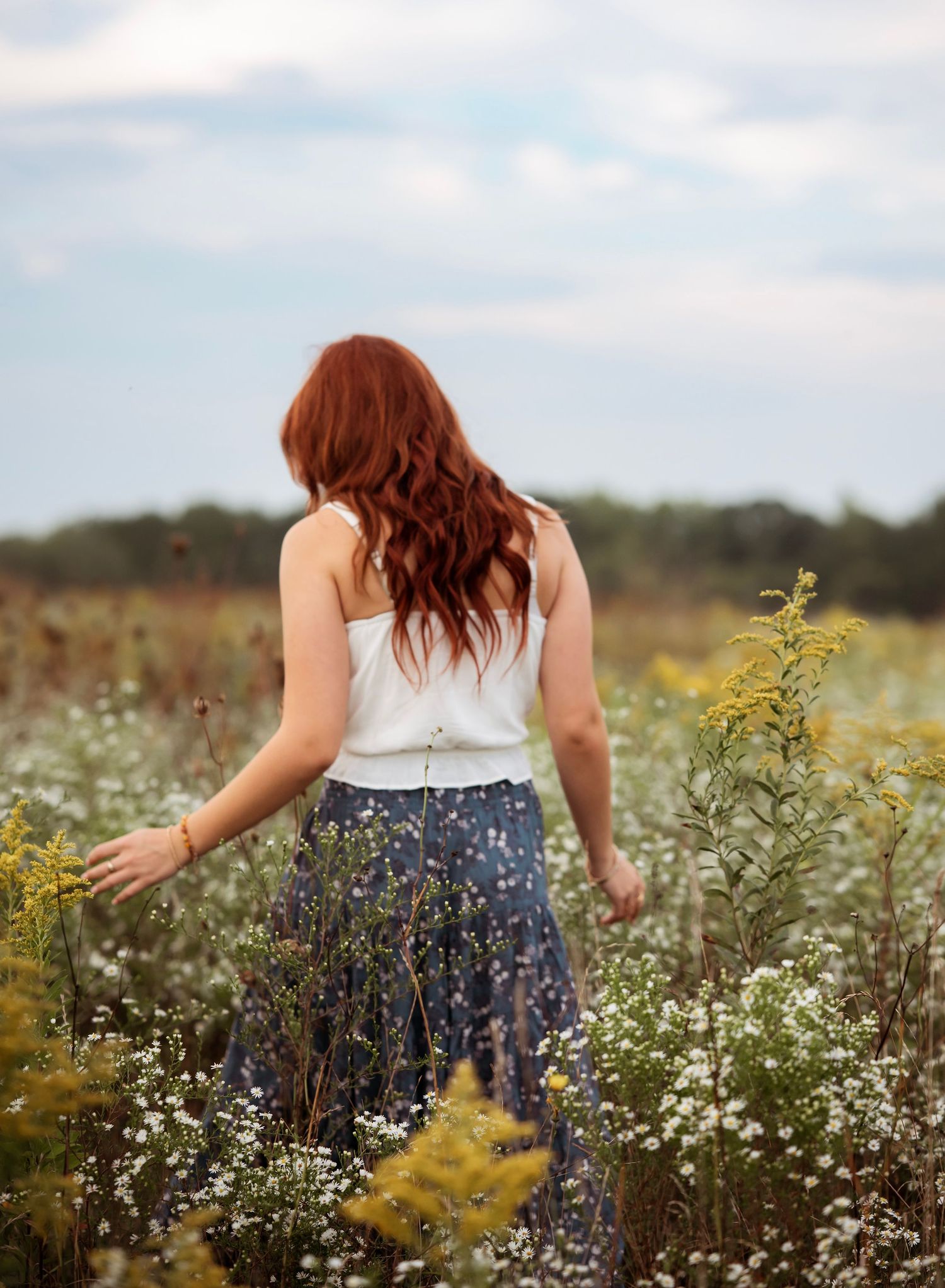 A person in a floral skirt walks through a dreamy meadow filled with delicate white wildflowers and golden grasses.