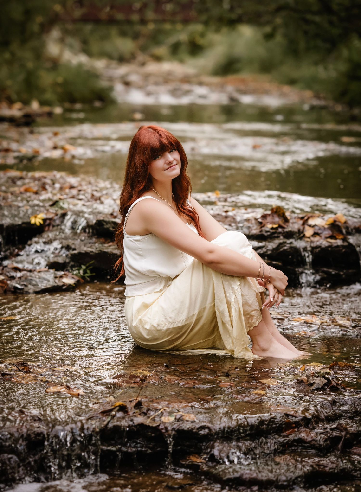 A dreamy portrait in a white dress sitting on rocks by a flowing stream in soft natural lighting.