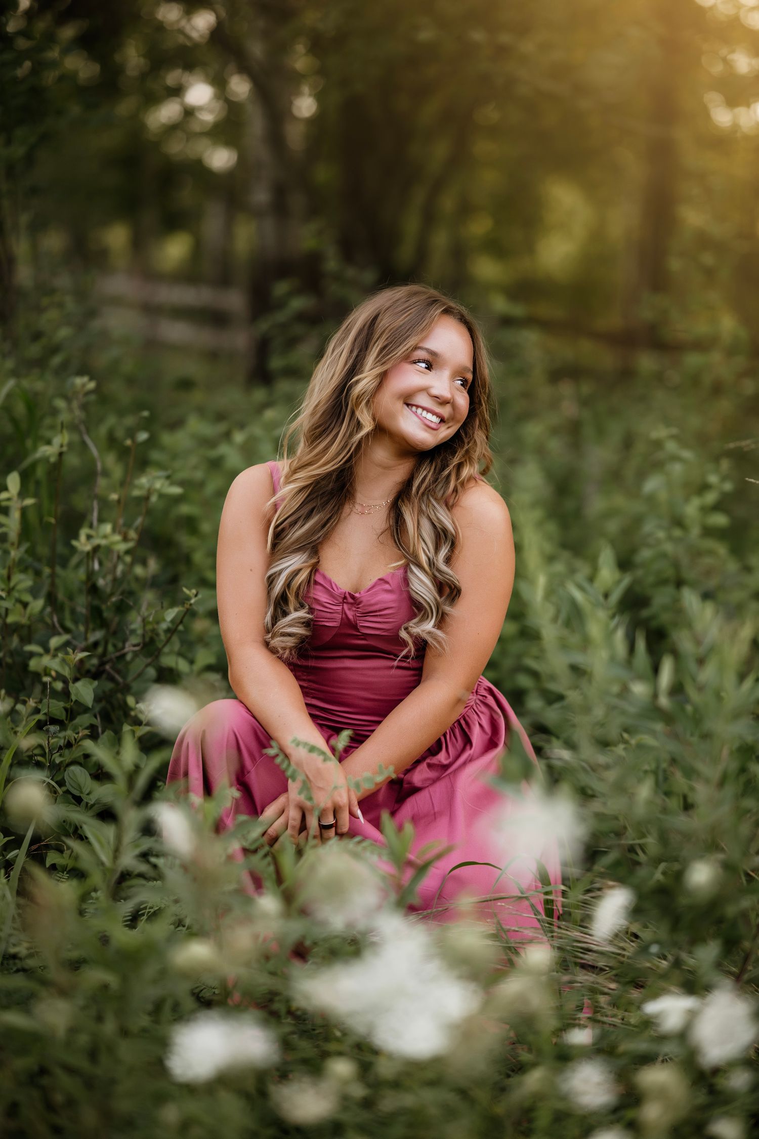 Young woman in pink dress sitting among wildflowers and lush greenery, smiling in golden hour sunlight.