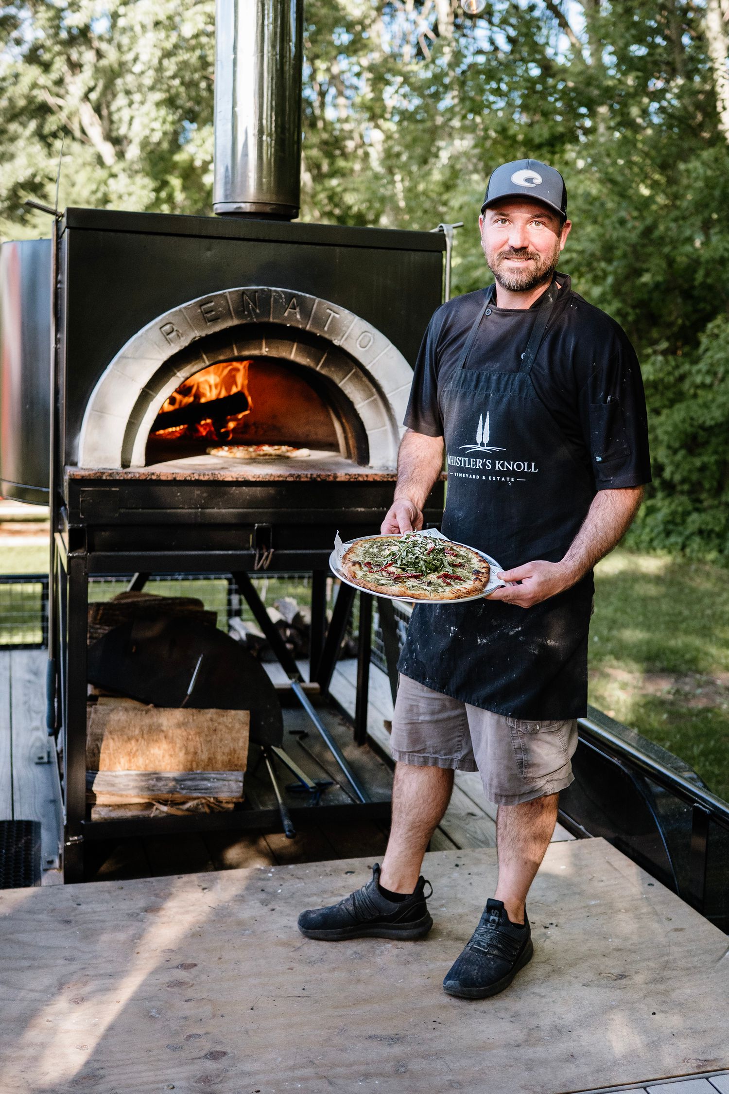 Chef holding a freshly baked pizza in front of a wood-fired oven outdoors, wearing a black apron and cap.