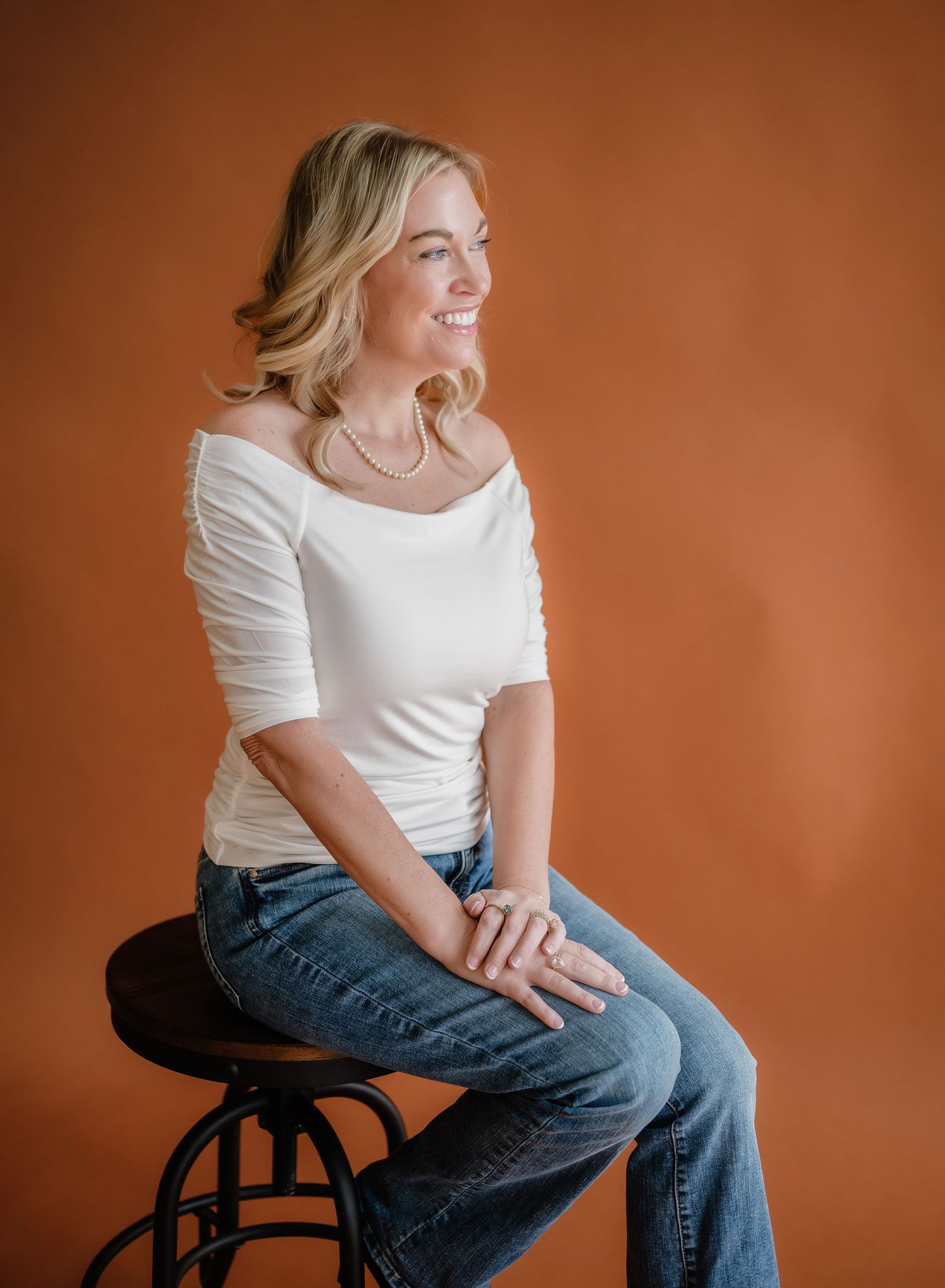 Portrait of someone wearing a white top and jeans while sitting casually on a black stool against an orange backdrop.