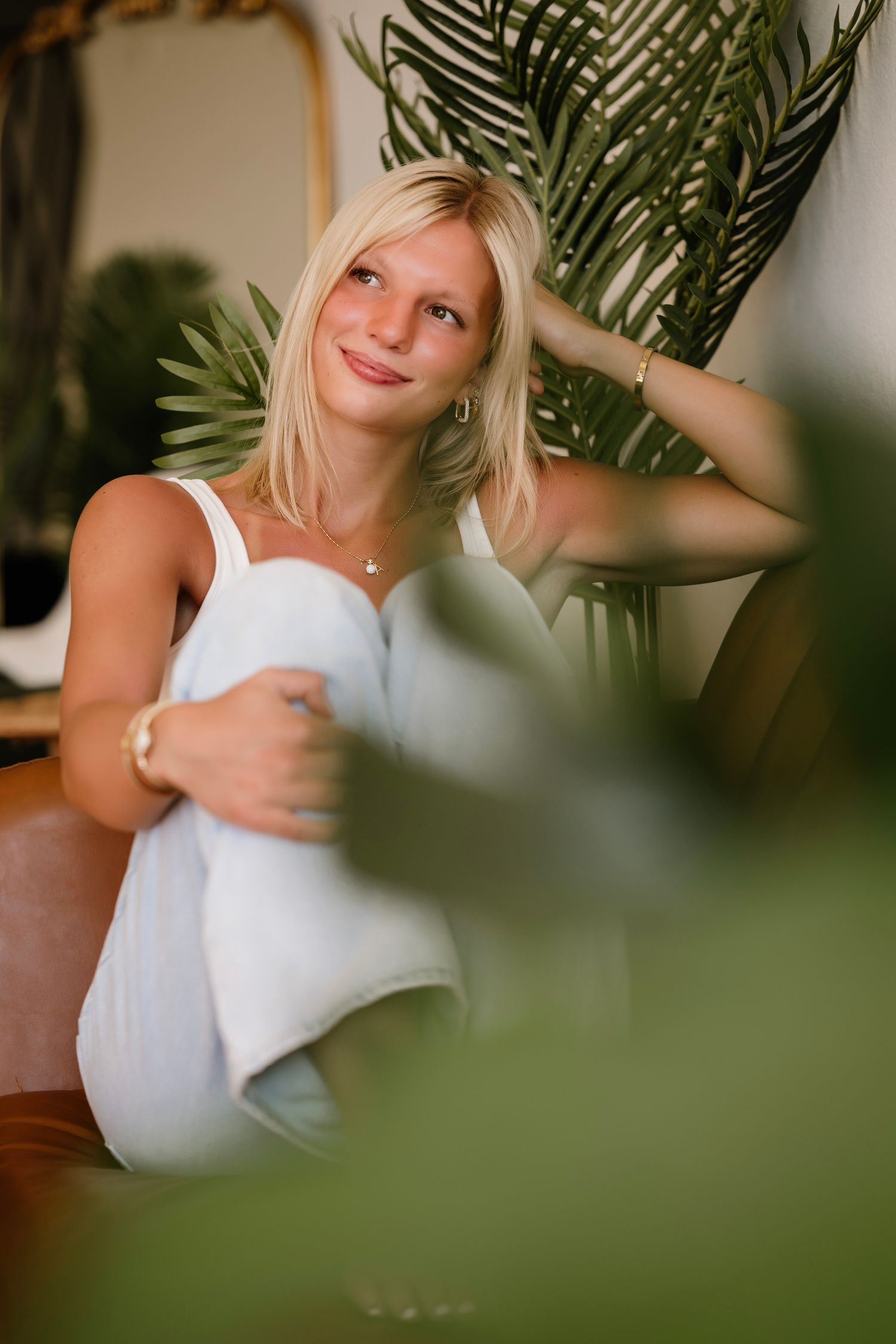 Blonde woman in white outfit relaxing among tropical plants, smiling softly in a cozy indoor setting.