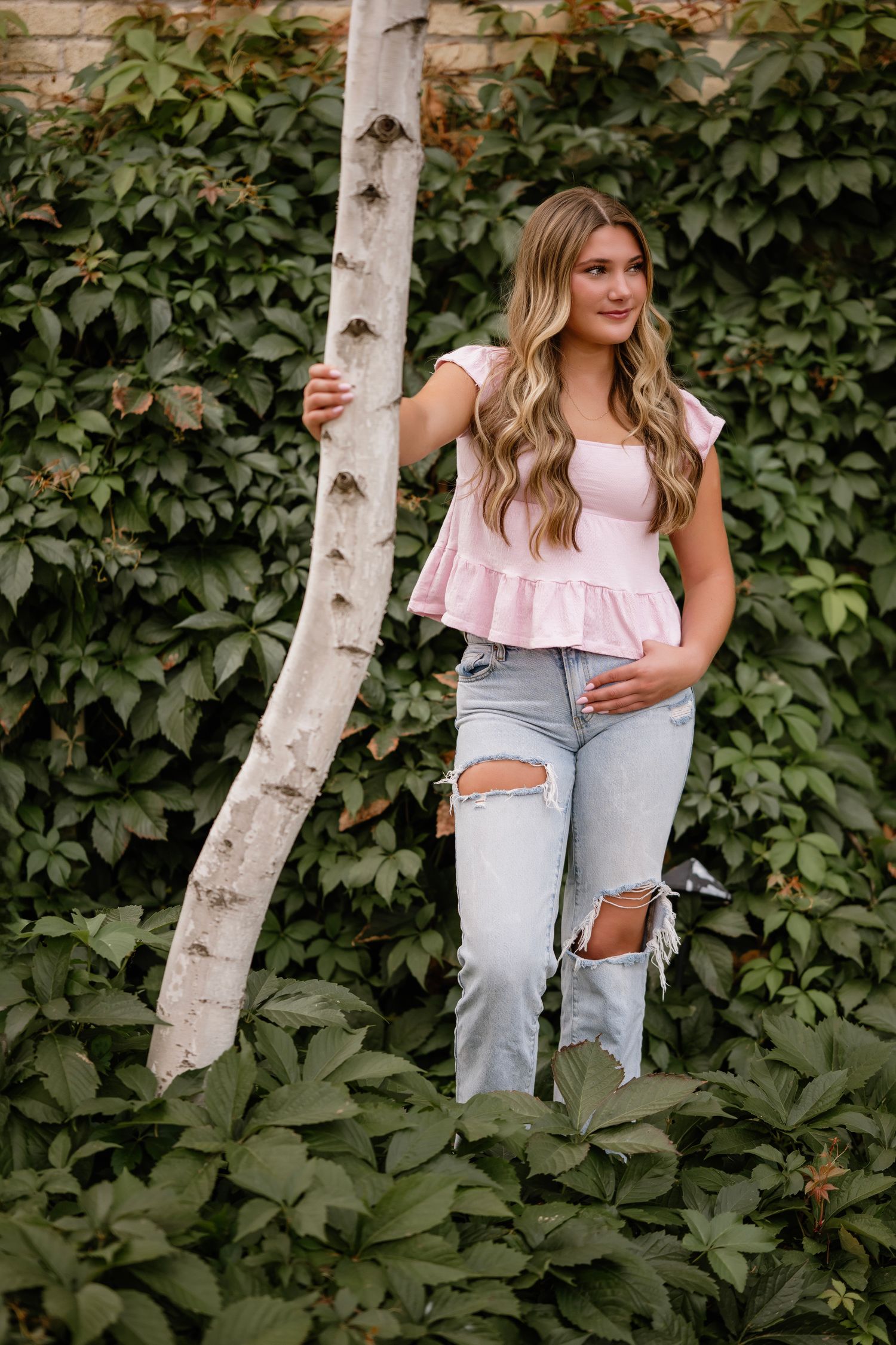 Young woman in pink ruffled top and ripped jeans poses by a birch tree surrounded by lush green foliage.