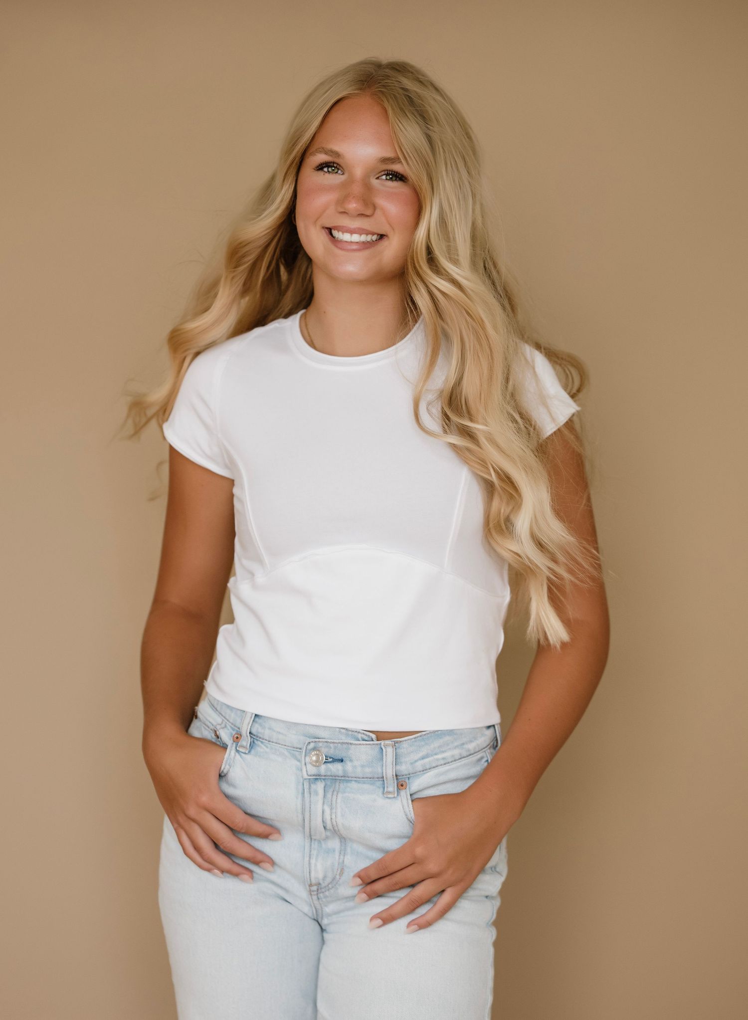 Smiling blonde teen girl in white crop top and light jeans posing against a tan backdrop.