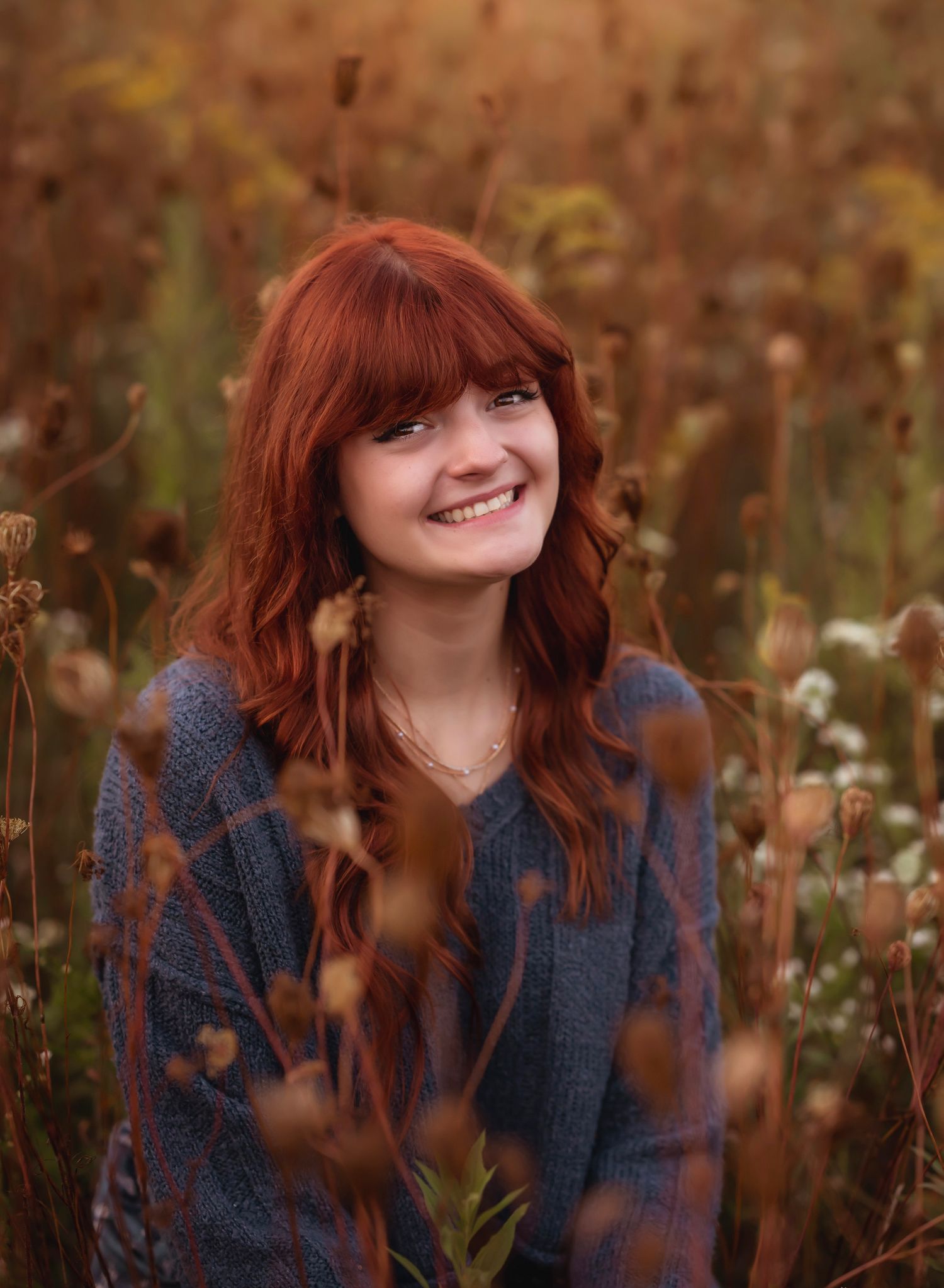 A person with red hair and bangs smiles joyfully while sitting in a field of autumn wildflowers at golden hour.