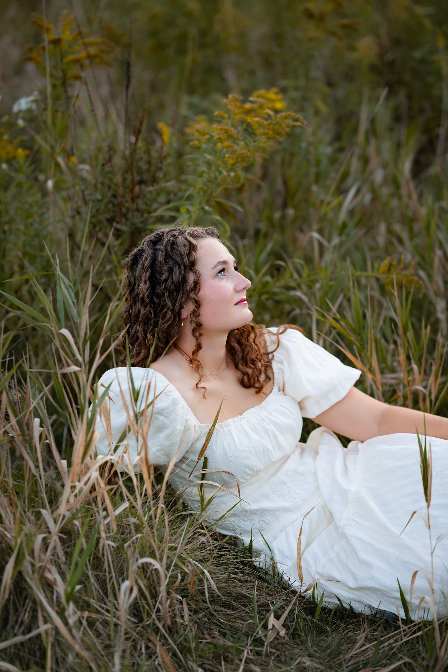 A woman in a white dress reclines in tall grass with yellow wildflowers in the background during golden hour.