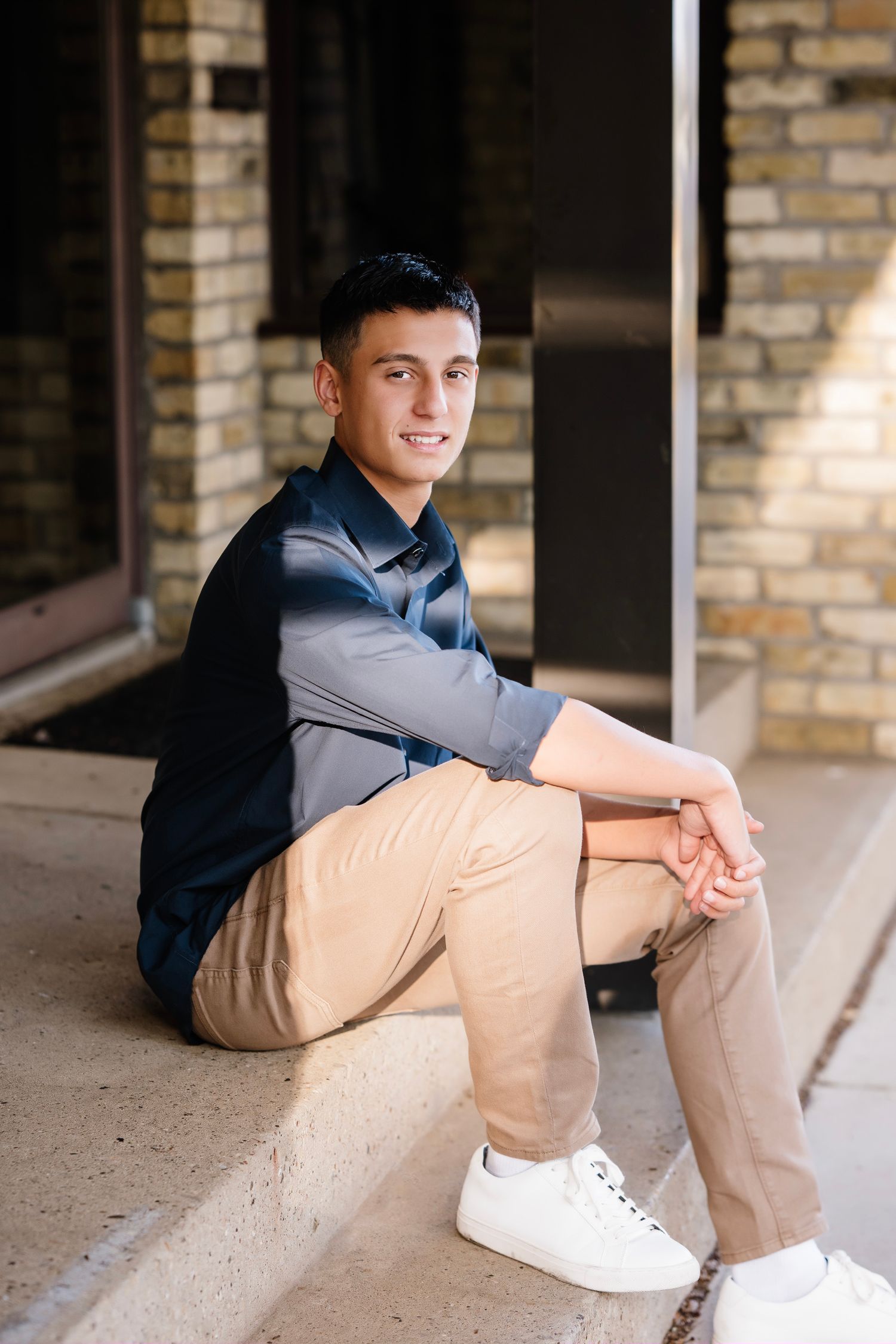 A person in a gray shirt and khaki pants sits casually on a brick porch with white sneakers.