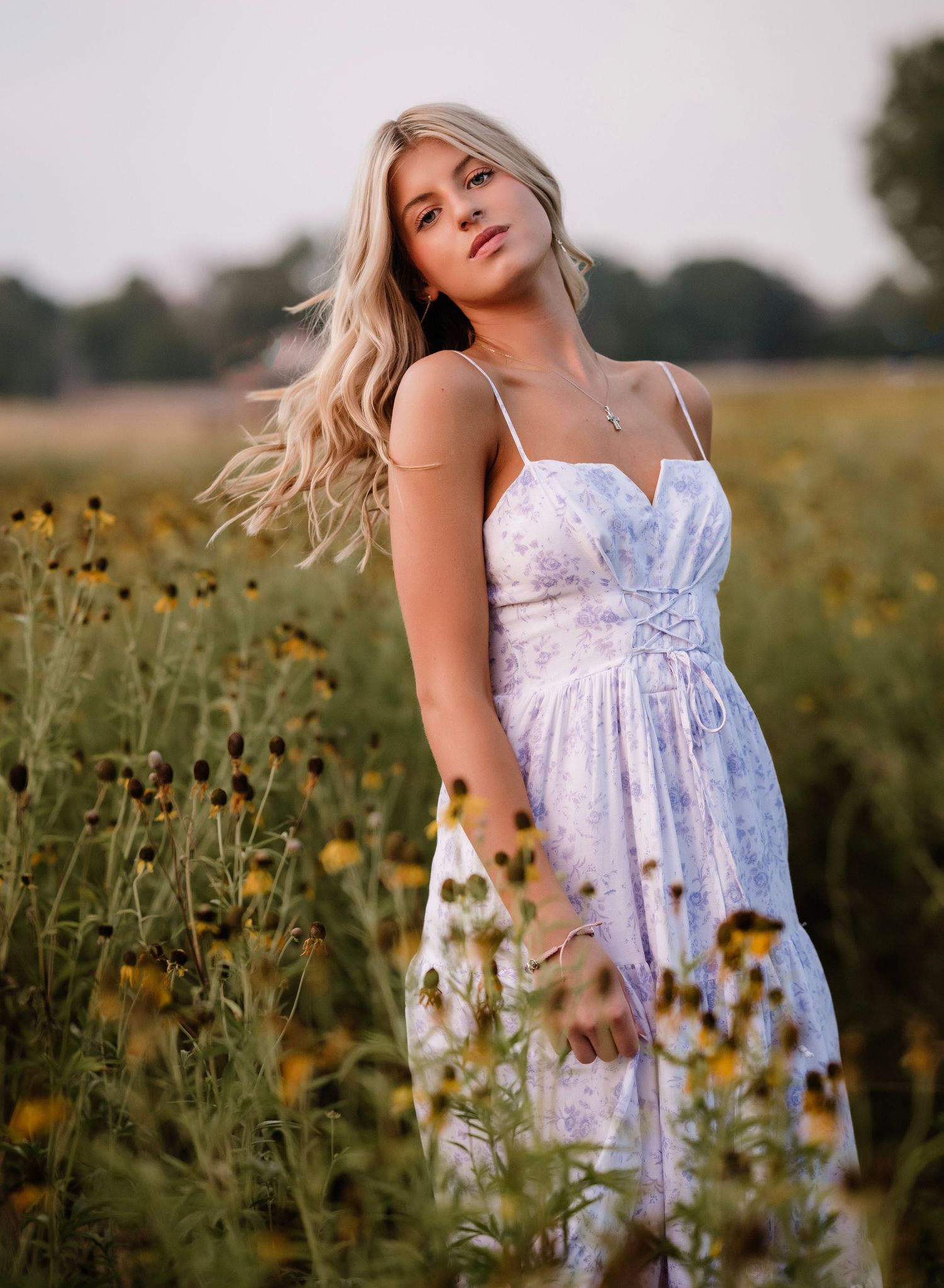 A person in a white floral dress poses in a field of yellow wildflowers during golden hour sunset.