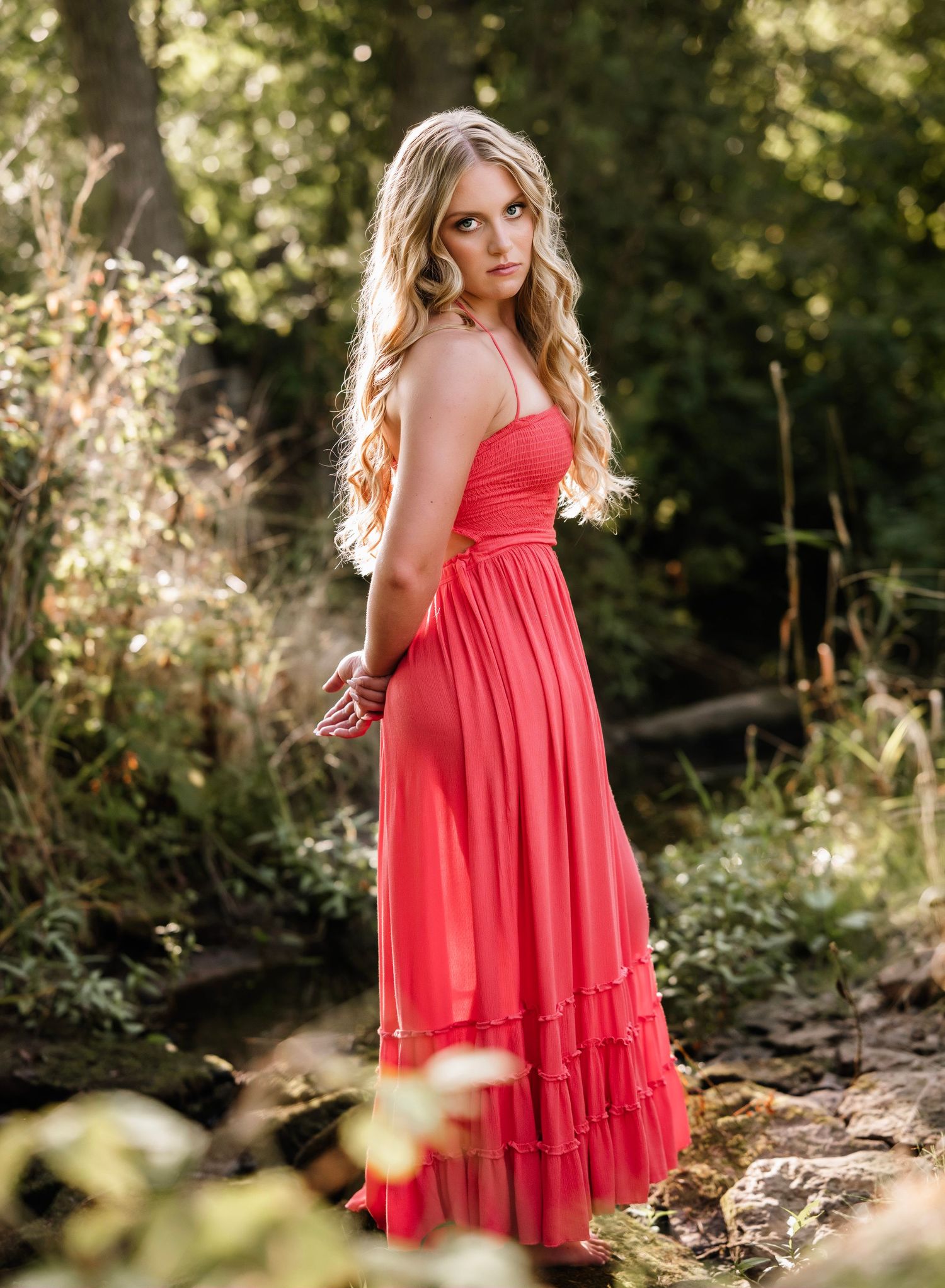 A flowing coral maxi dress with tiered ruffles is worn in a sunlit outdoor garden setting with natural greenery.