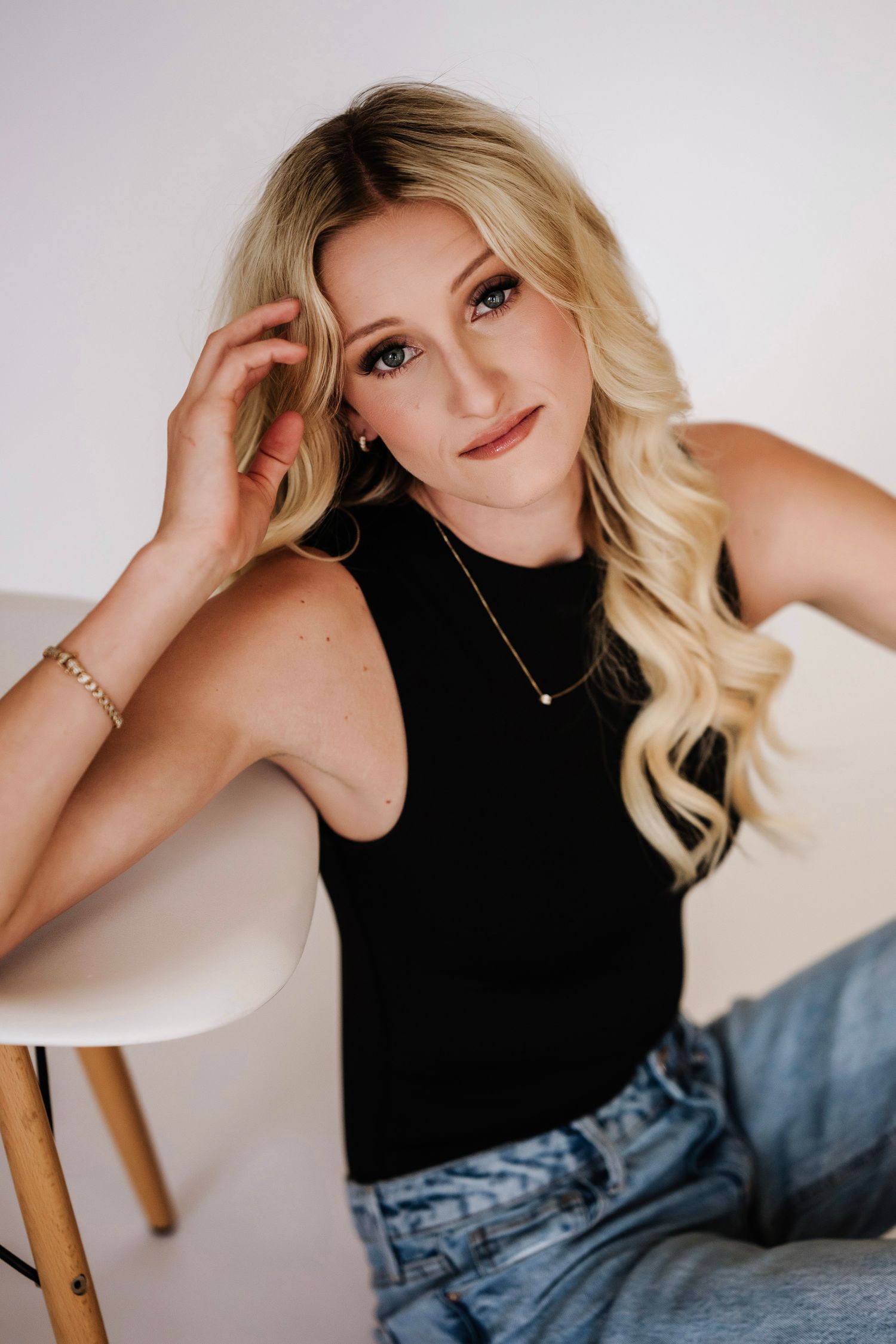 Blonde woman in black sleeveless top and jeans, wearing gold jewelry, posing casually beside a white chair.