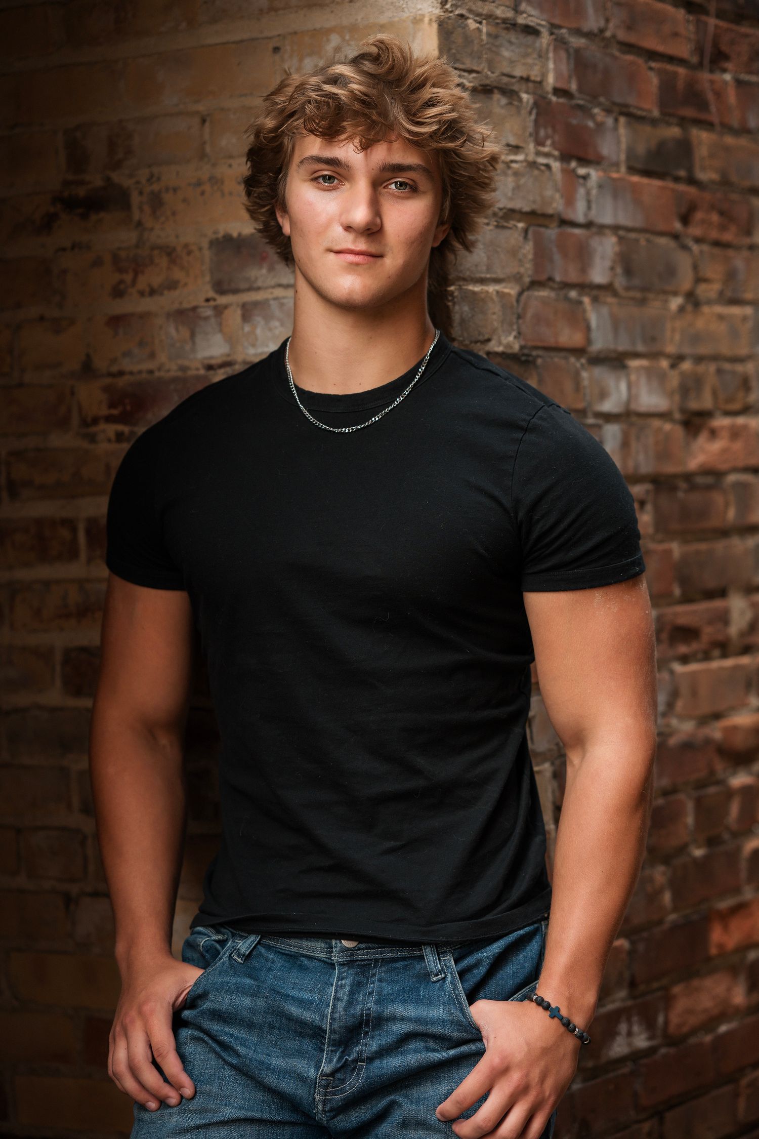 Young person in a black t-shirt and jeans poses against rustic brick wall background with natural lighting.