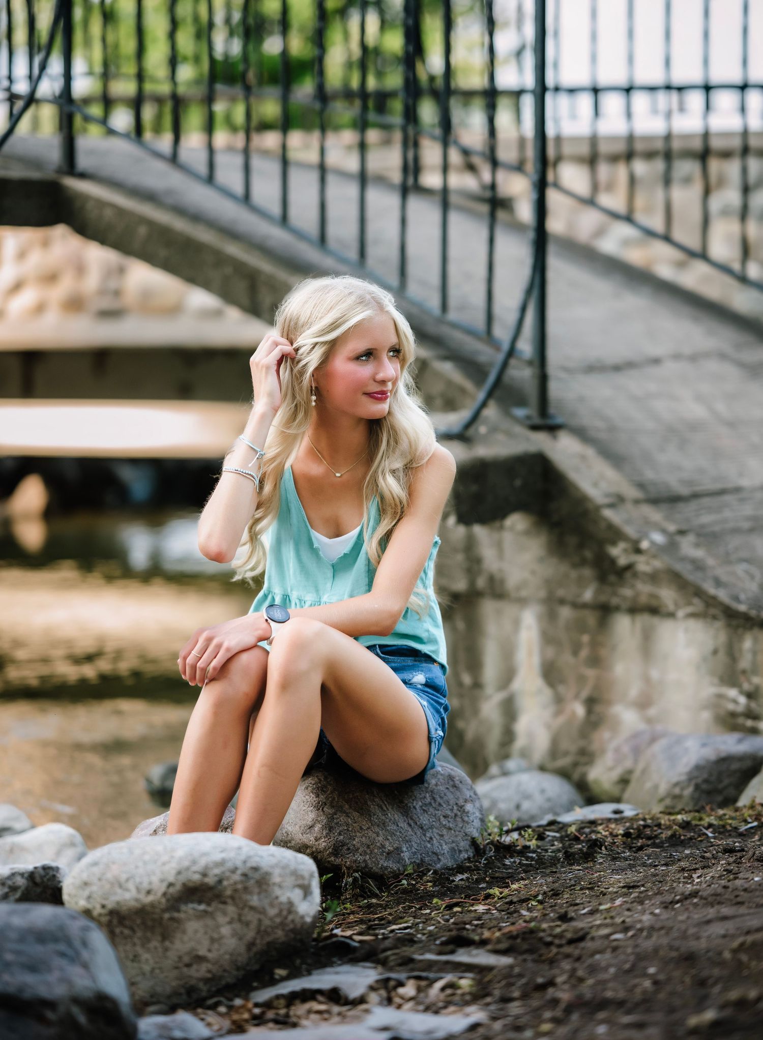 A person in a blue tank top sits on a rock by a stream beneath a bridge on a summer day.