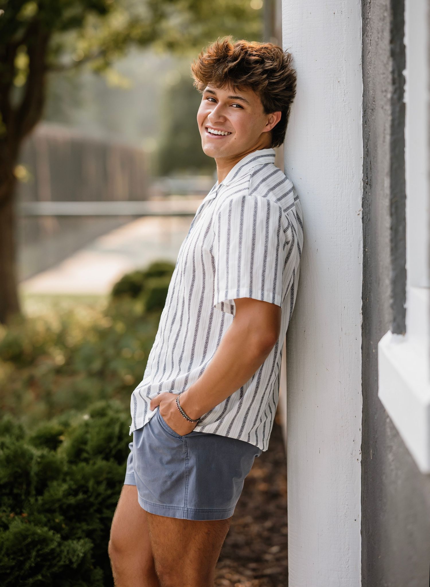 A person wearing a striped shirt and shorts poses playfully against a white column in an outdoor setting.