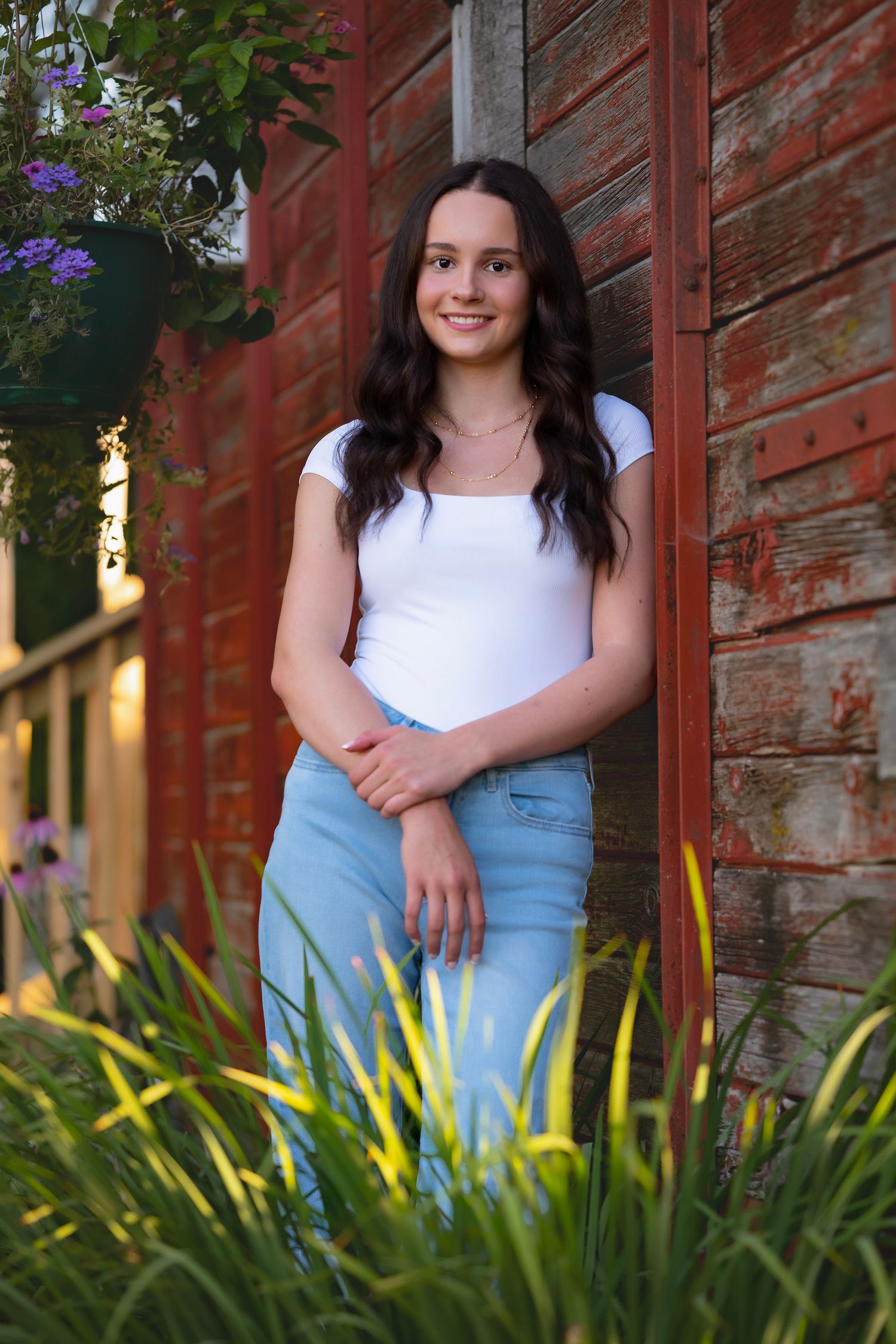 Young woman in white top and jeans smiling outdoors by rustic red barn wall with lush green plants.