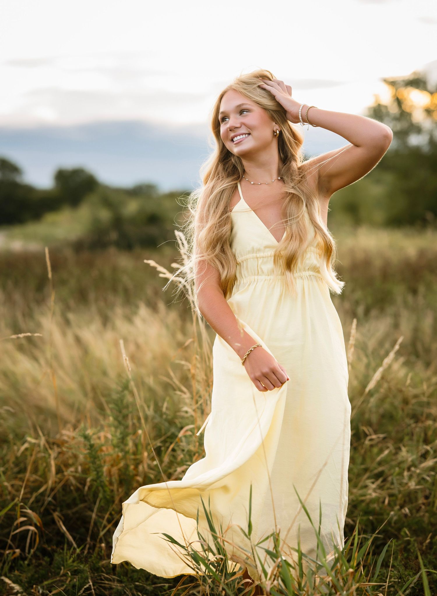 Young woman in flowing yellow dress smiling and walking through tall grass field at sunset.