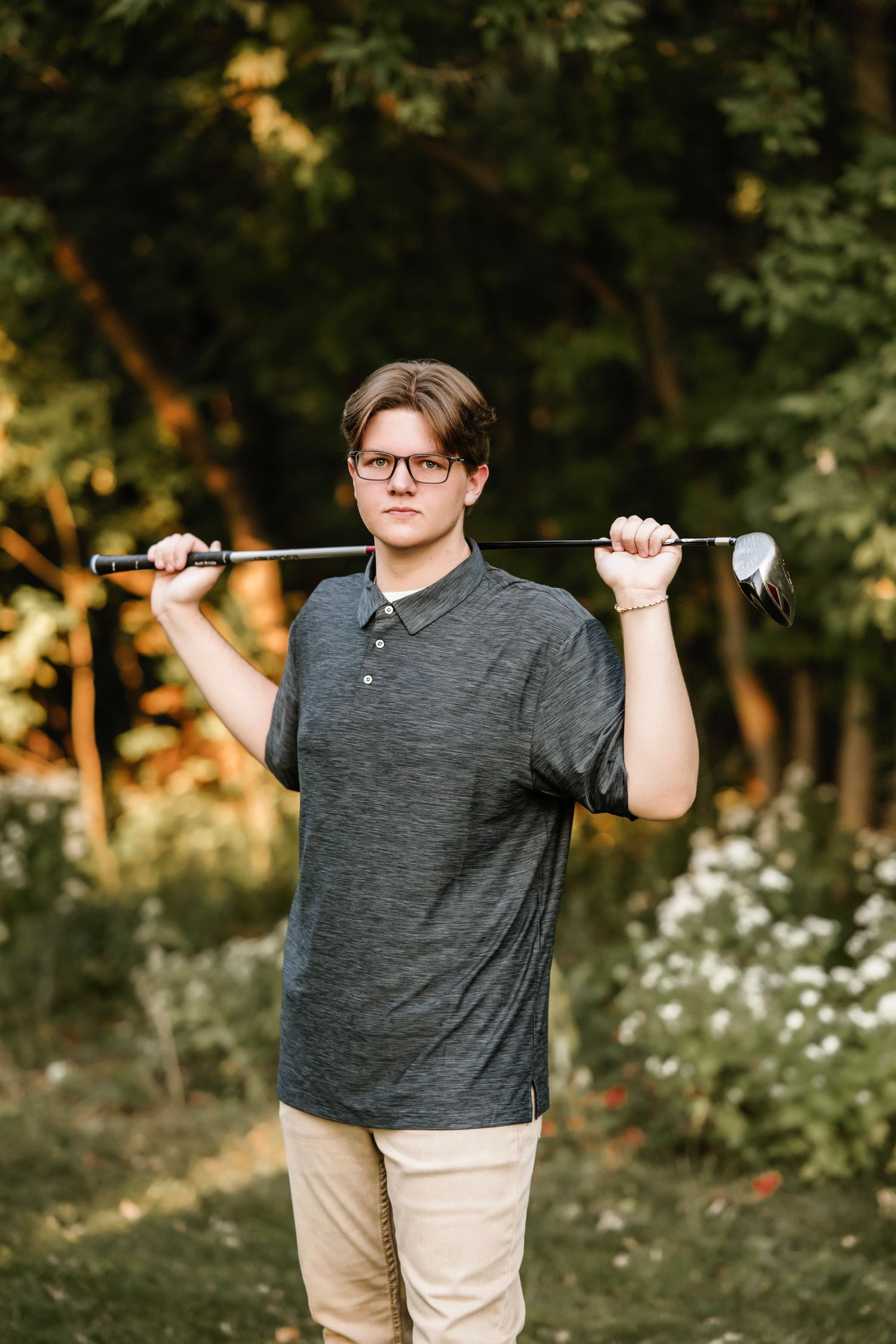 A golfer in a gray polo shirt and khaki pants holds a golf club across their shoulders in a sunlit outdoor setting.