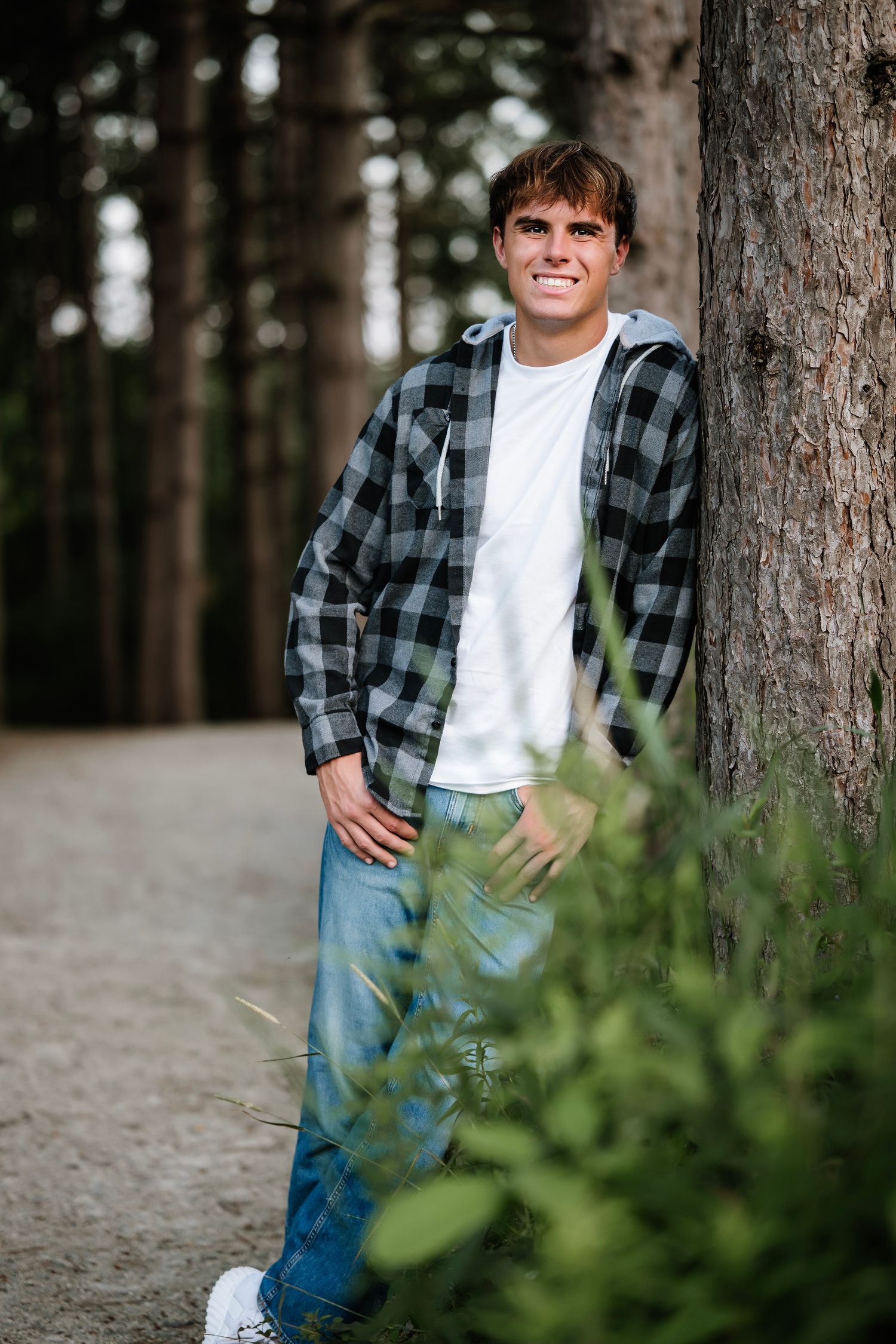 Casual outdoor portrait against pine trees showing someone in a plaid flannel shirt and jeans leaning on tree trunk.