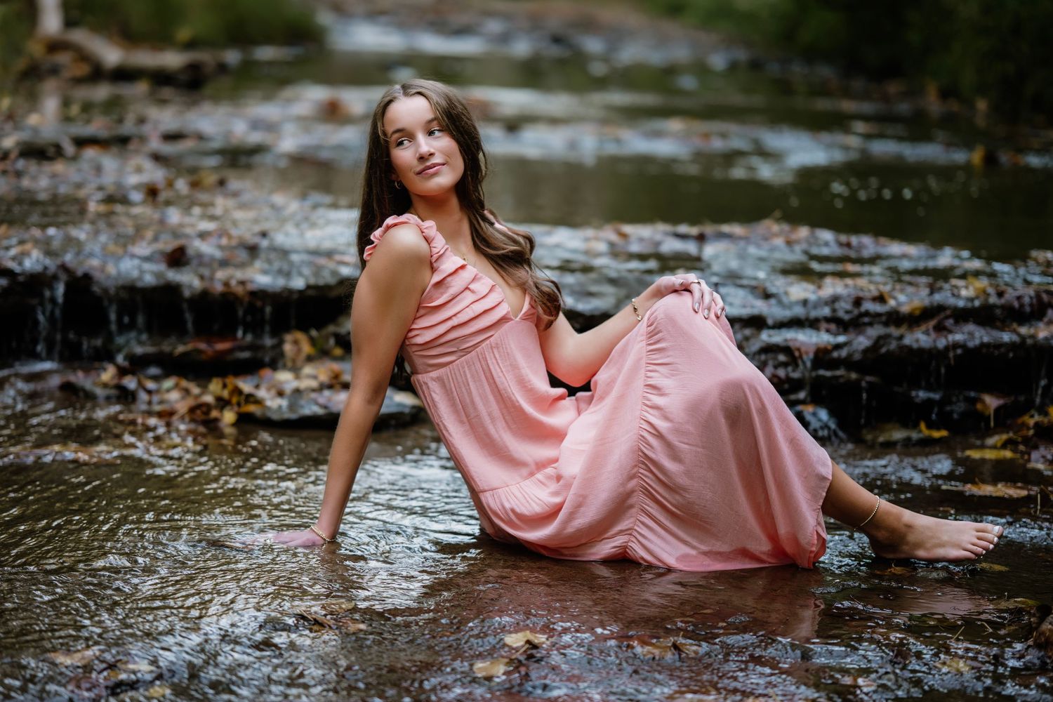A person in a flowing pink dress poses gracefully on wet stone steps surrounded by autumn leaves.