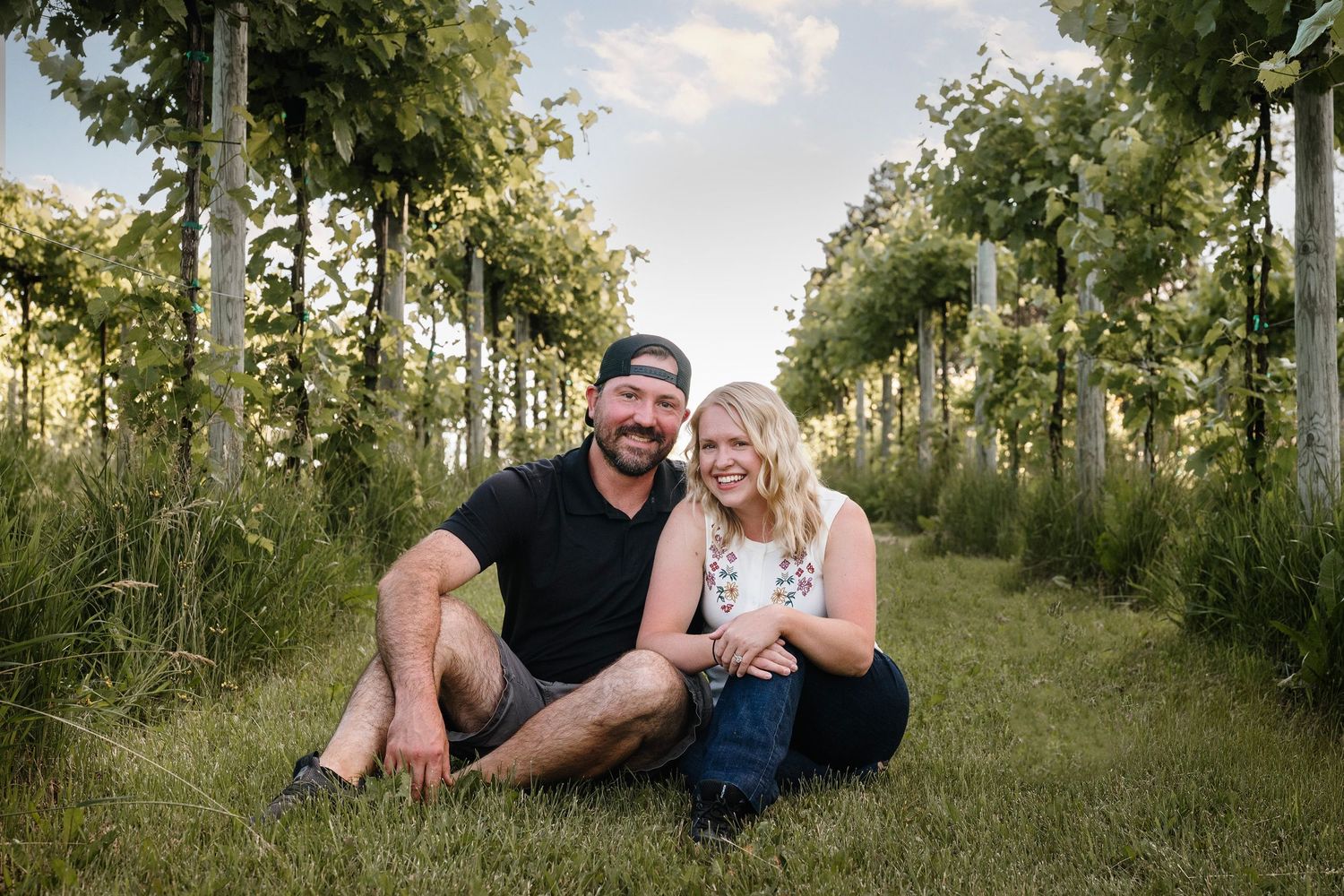 Couple sitting together on grass between vineyard rows, smiling outdoors during golden hour.