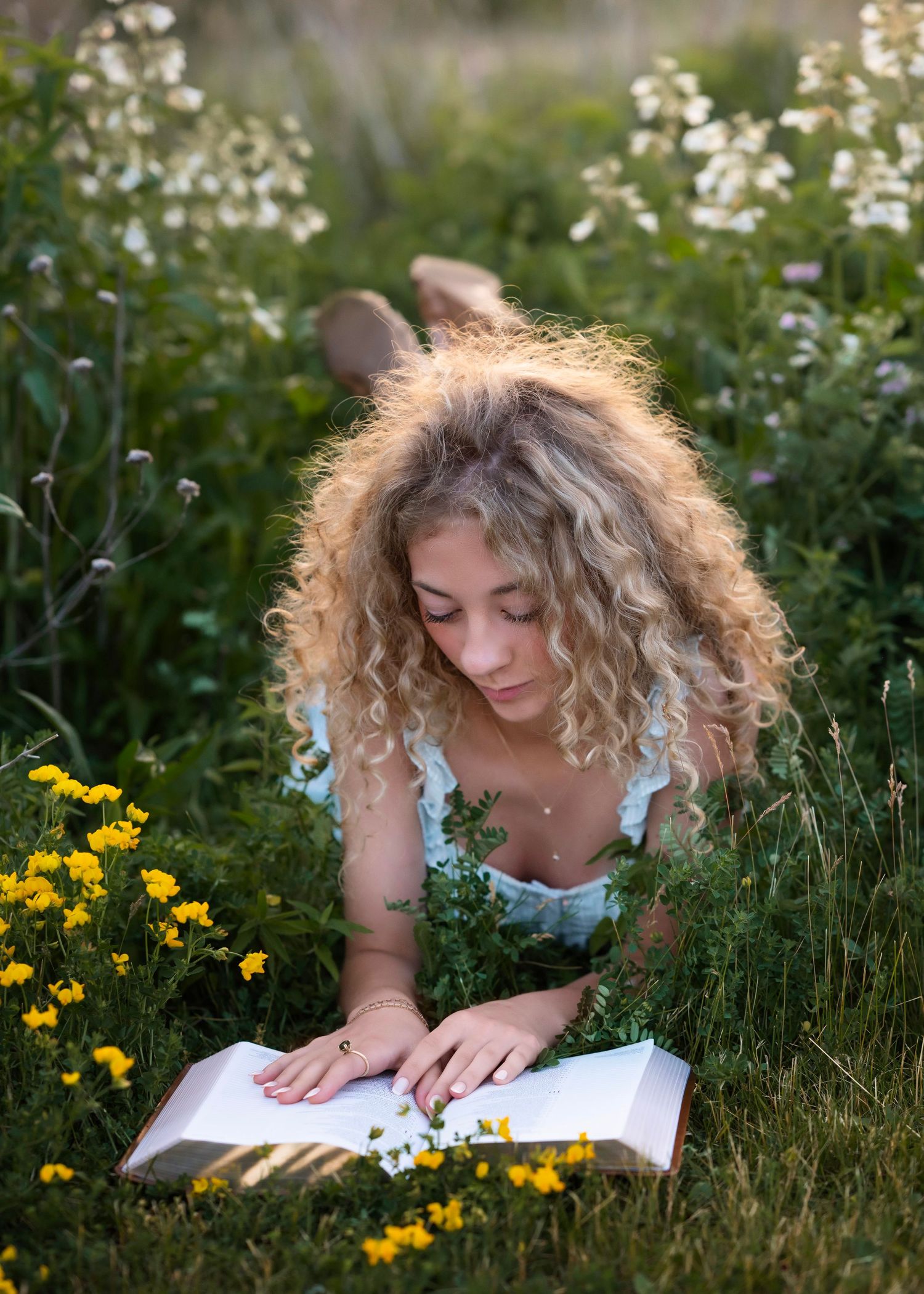 Young woman with curly blonde hair reading a book while lying in a field of yellow and white wildflowers.