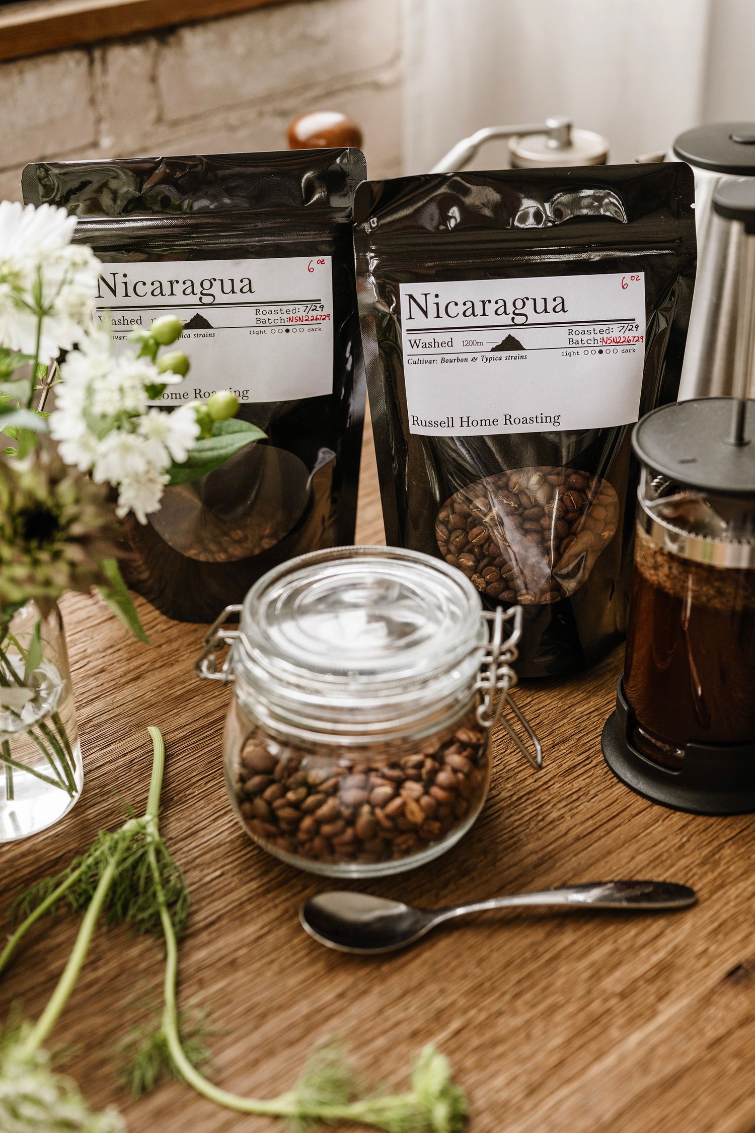 Two Nicaragua coffee bags by Russell Home Roasting displayed on a wooden table with a glass jar of coffee beans and French press.