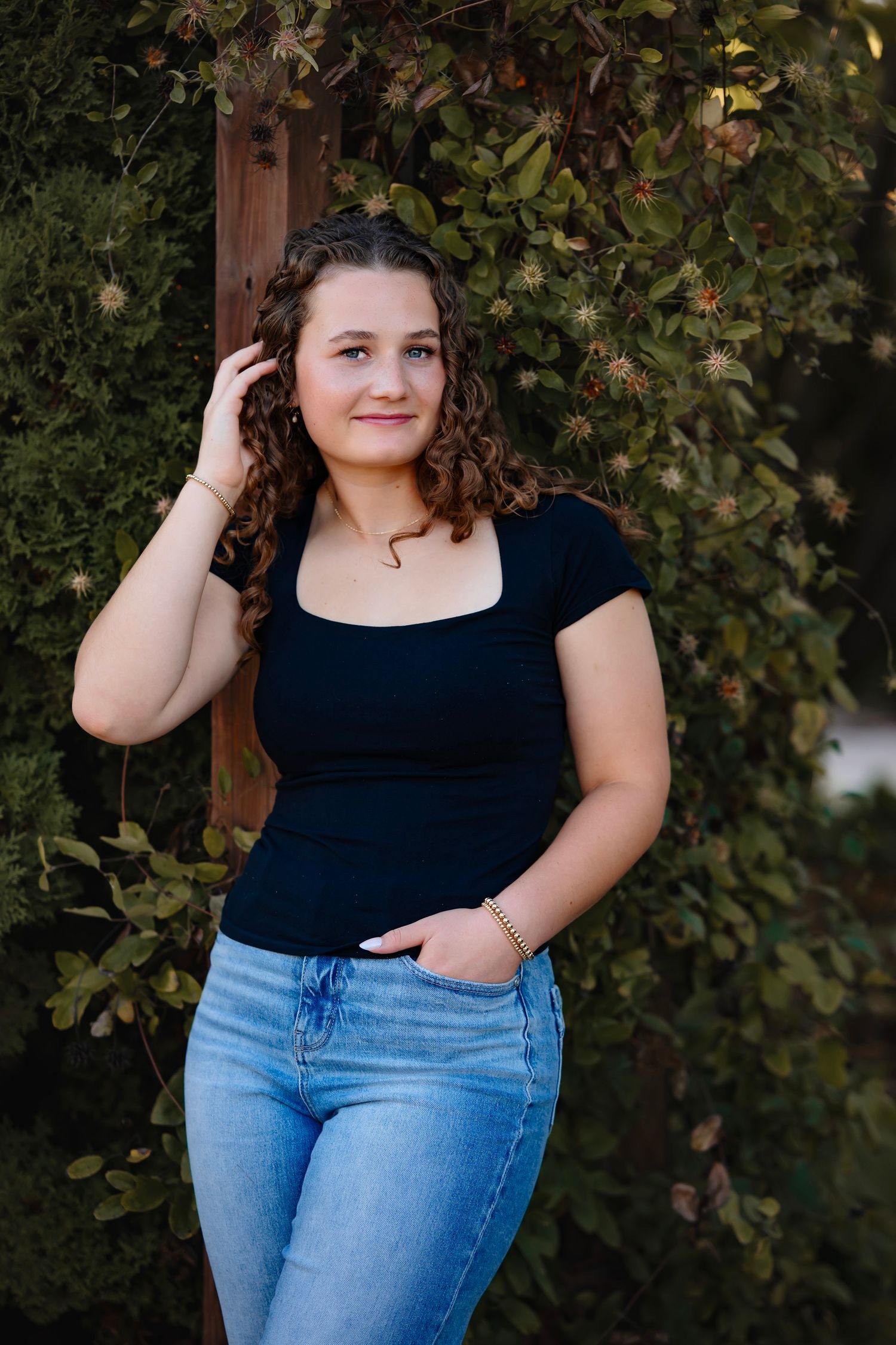 A person in a black top and blue jeans poses outdoors against a leafy green backdrop in natural lighting.