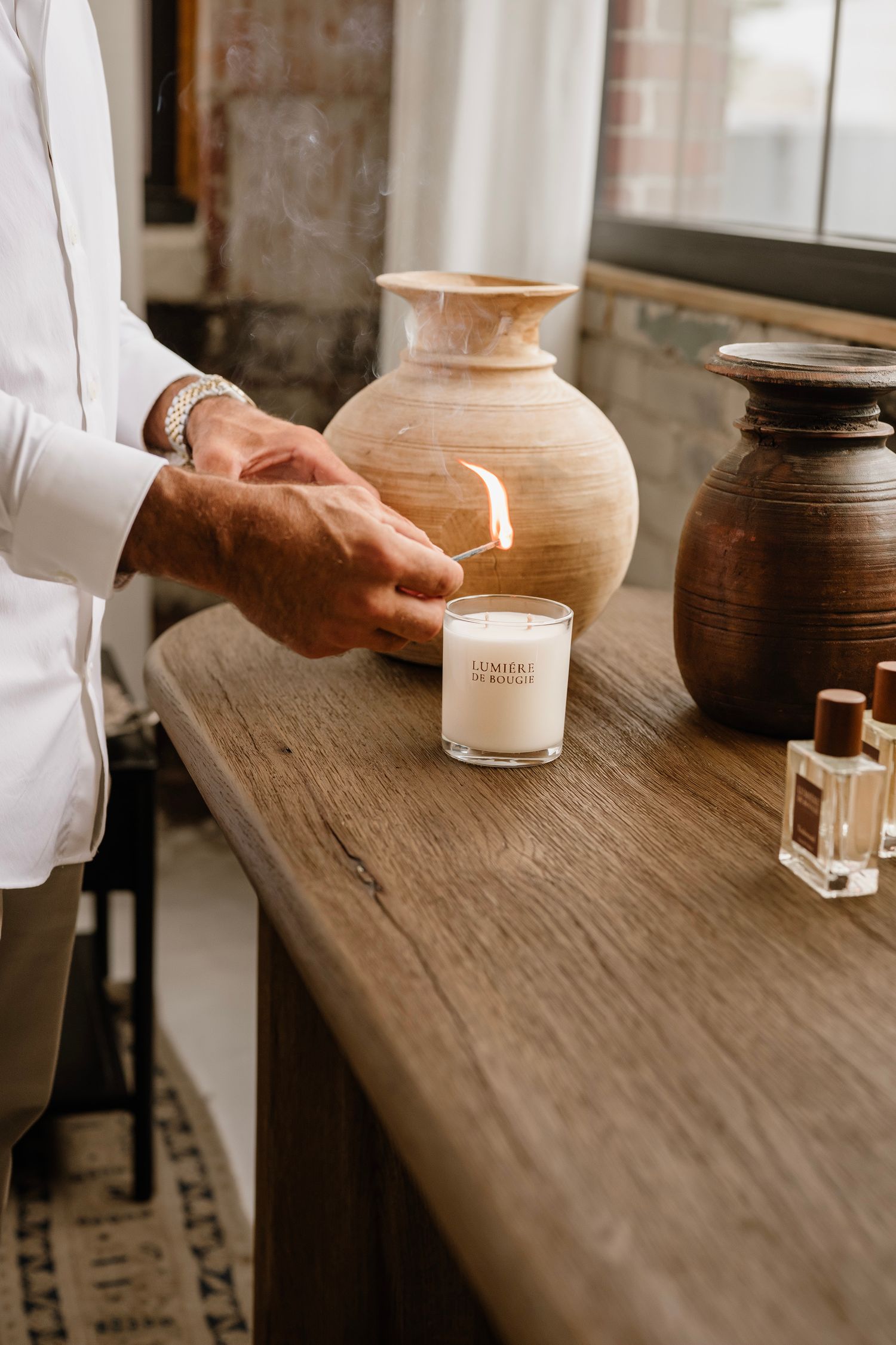 A person lighting a white candle on a rustic wooden console table with ceramic vases and perfume bottles.