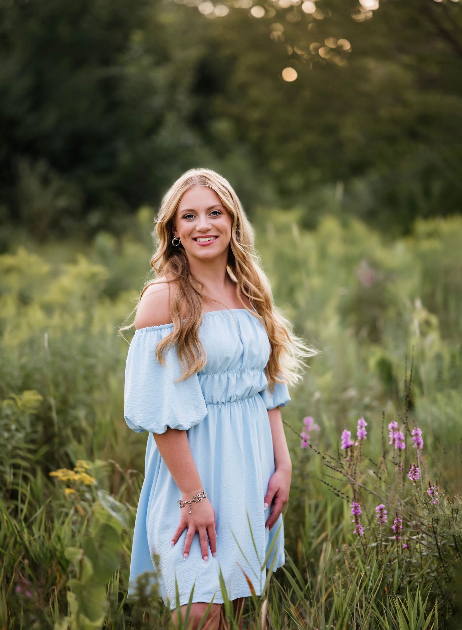 Young blonde woman in light blue off-shoulder dress smiling in a wildflower meadow during golden hour.