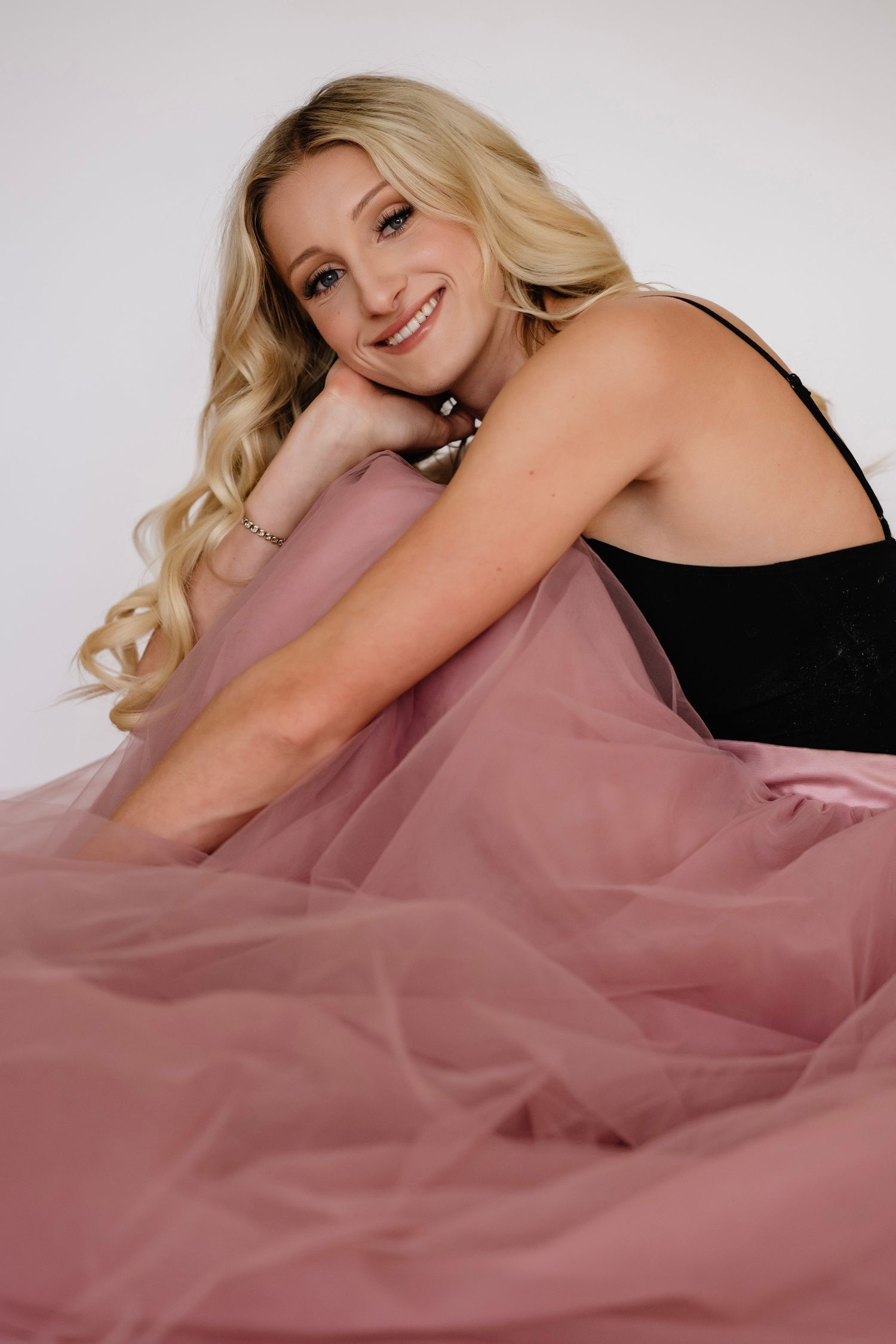 Smiling blonde woman in black top and pink tulle skirt posing gracefully on white studio background.