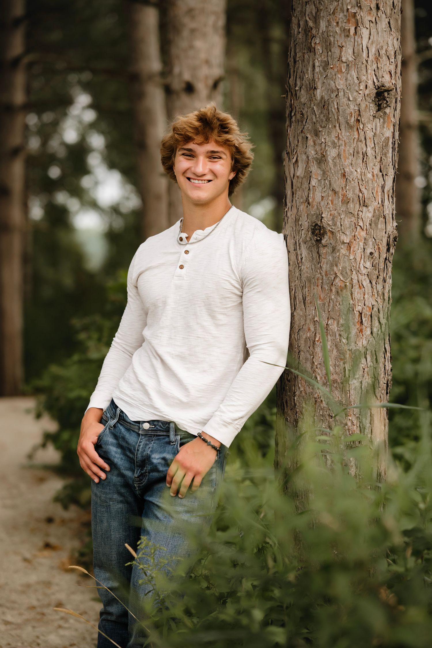 Person in white henley shirt and blue jeans poses casually against trees in outdoor portrait session.