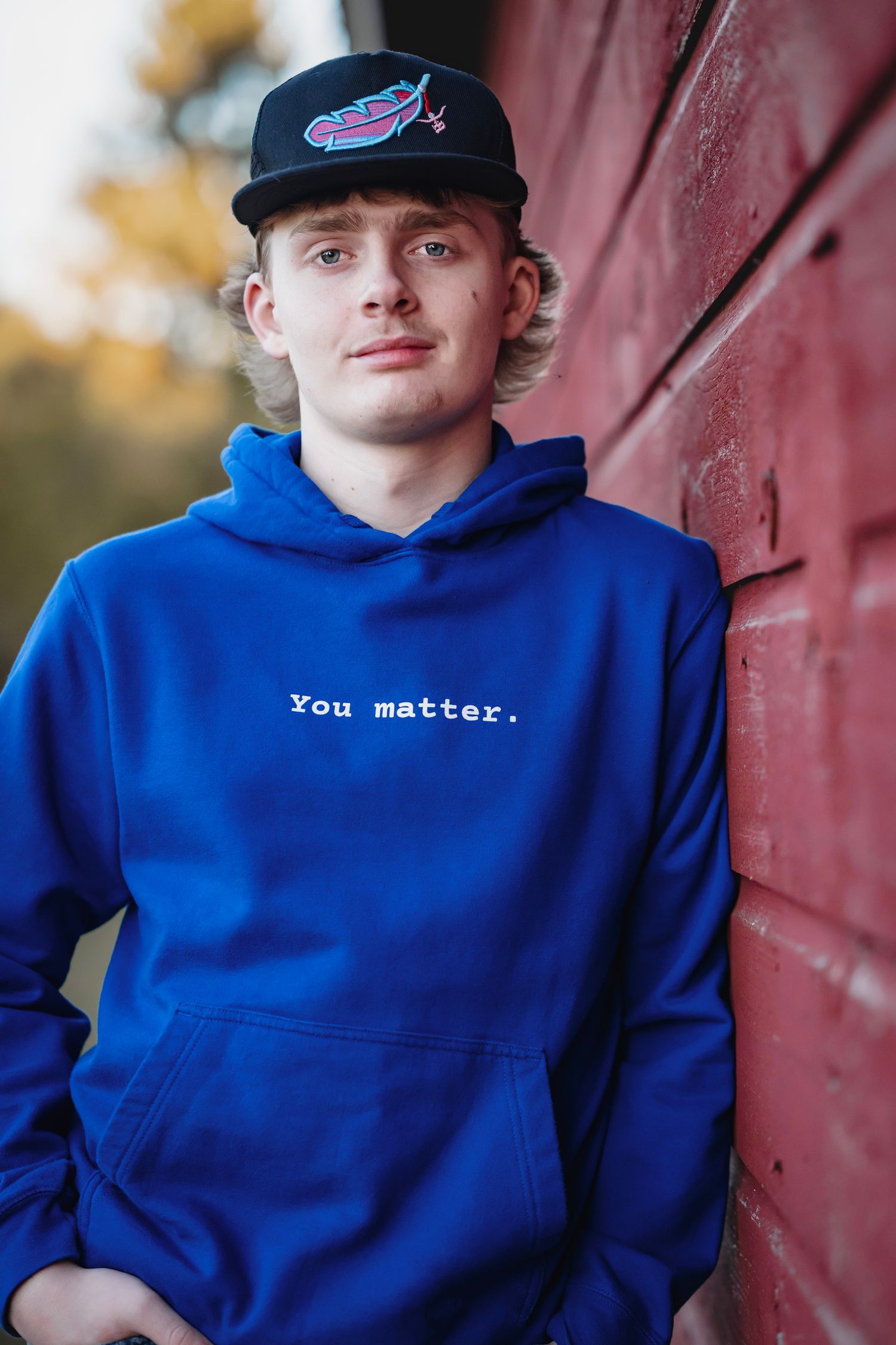 A person wearing a bright blue hoodie with 'You matter' text stands against a red barn wall in outdoor portrait photos.