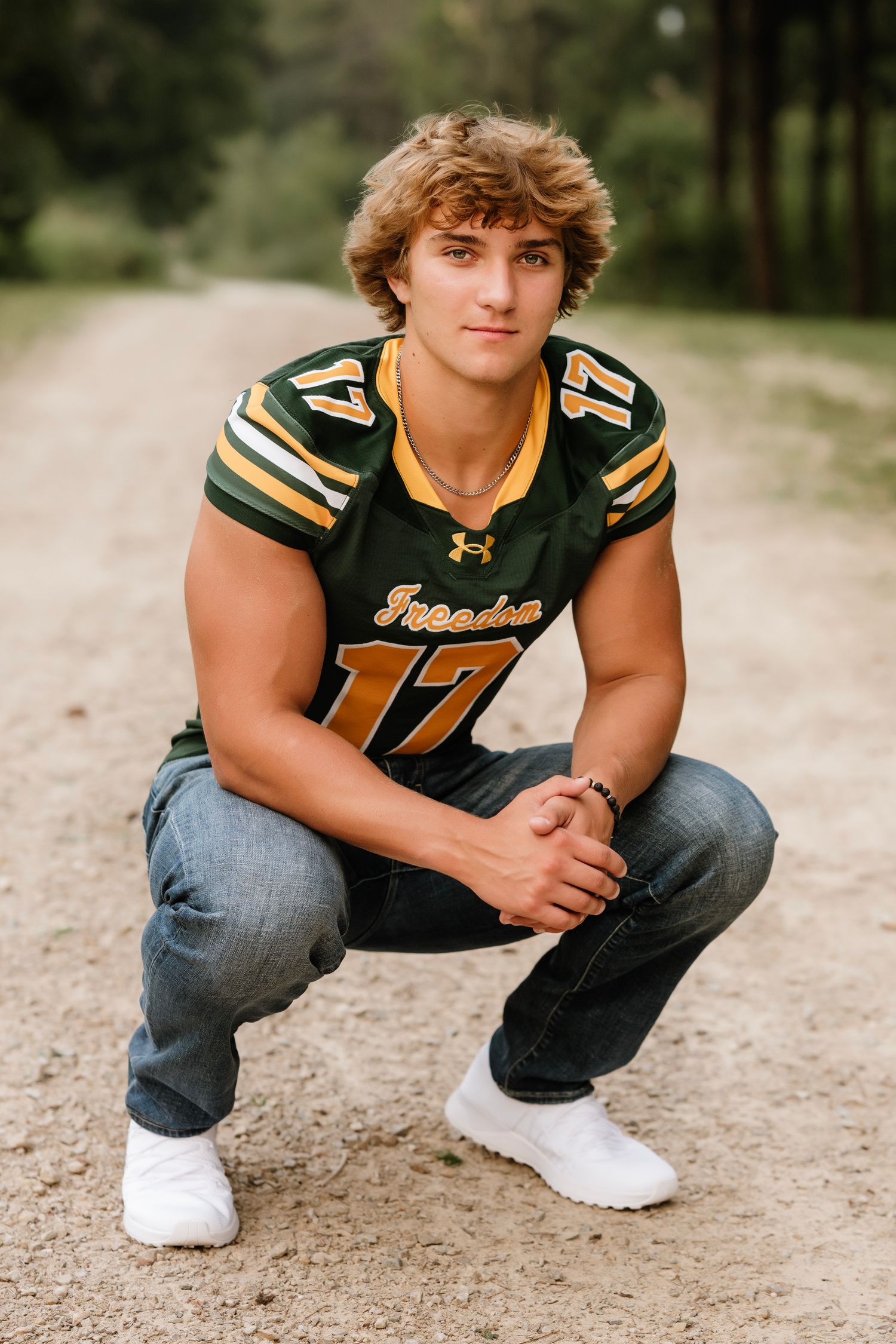 High school athlete in green and gold football jersey crouches on dirt path for senior portrait photos.