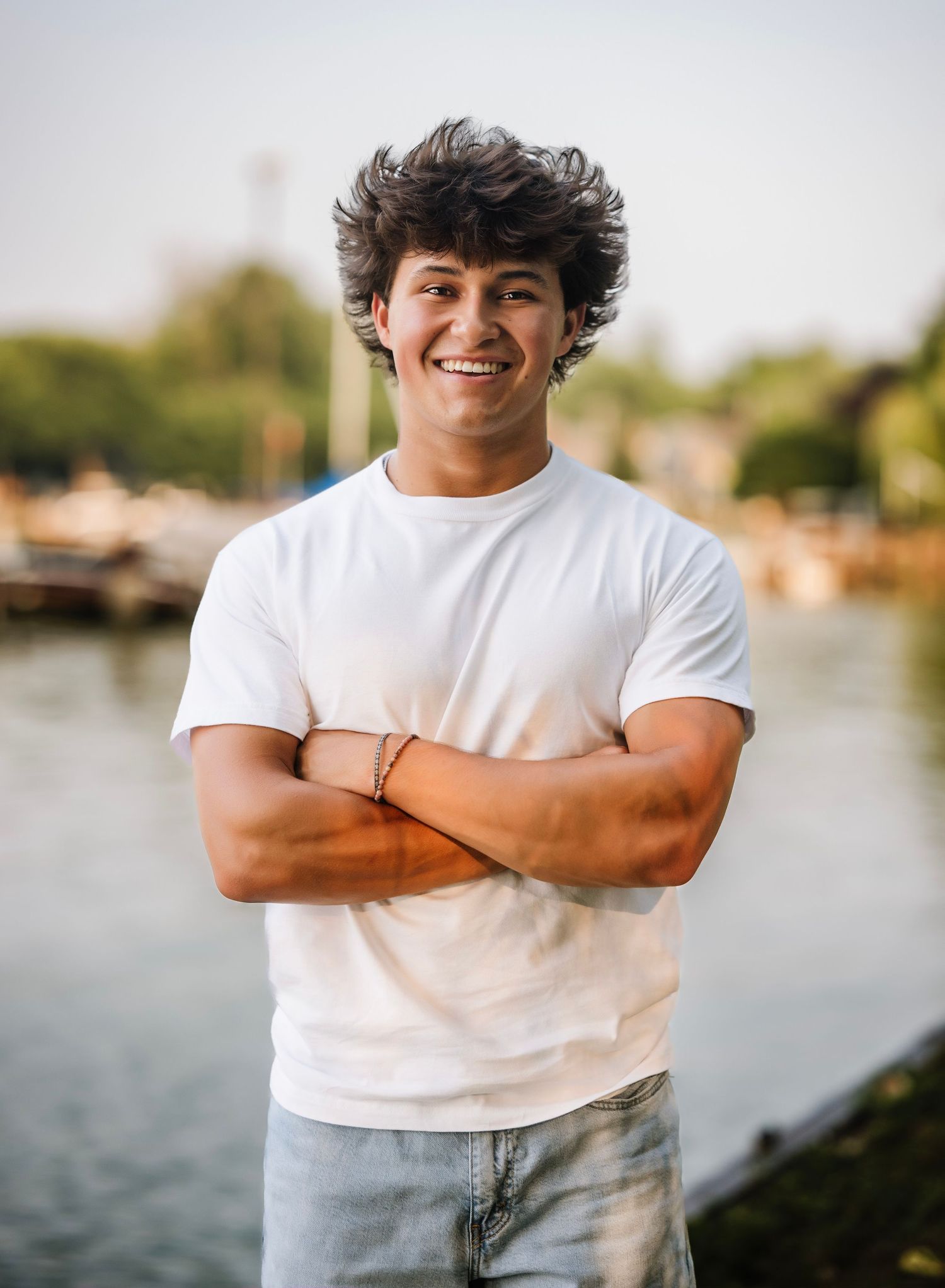 A person in a white t-shirt smiles and poses against a waterfront background during golden hour.