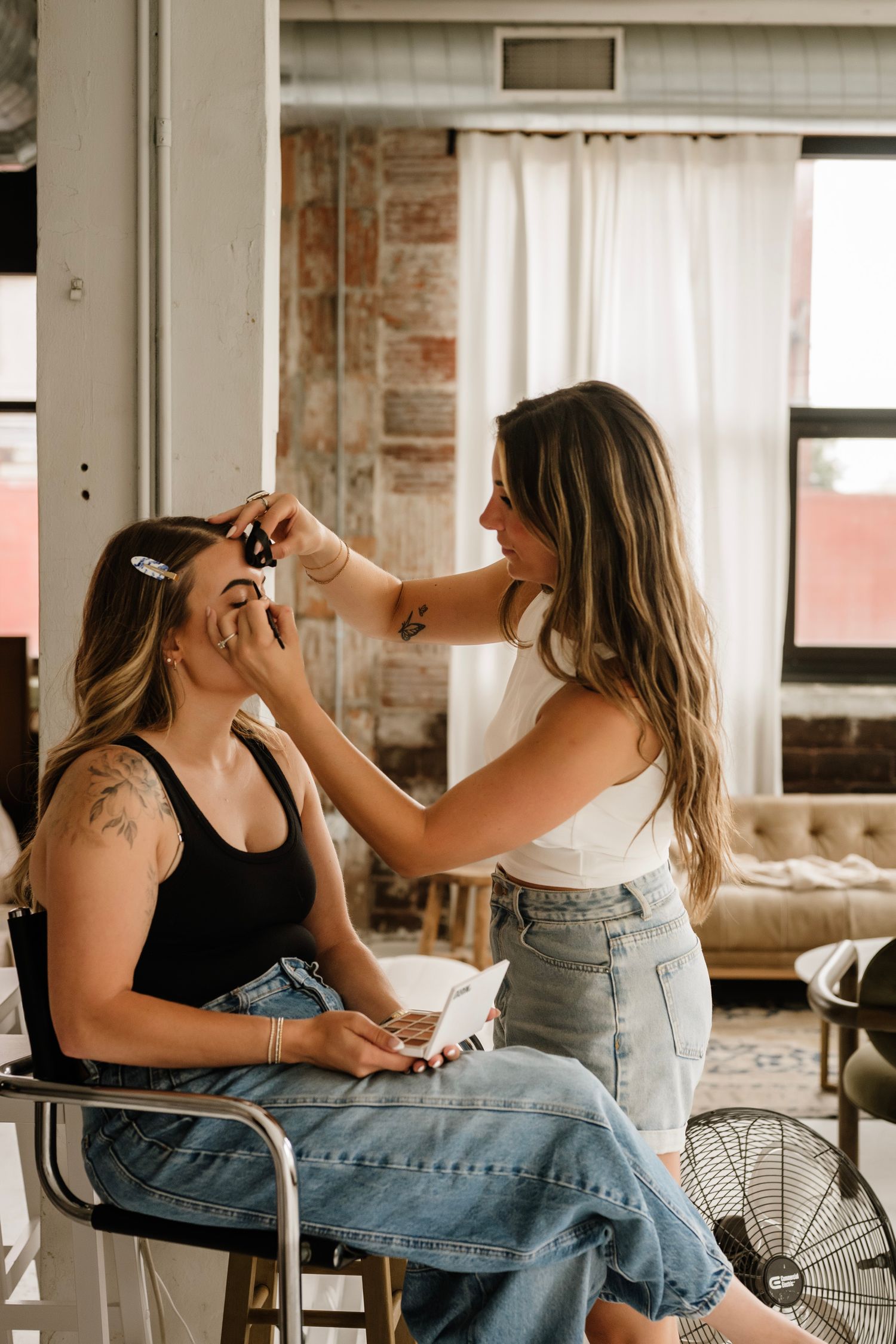 Makeup artist applying eye makeup to a seated woman in a bright studio with exposed brick walls.