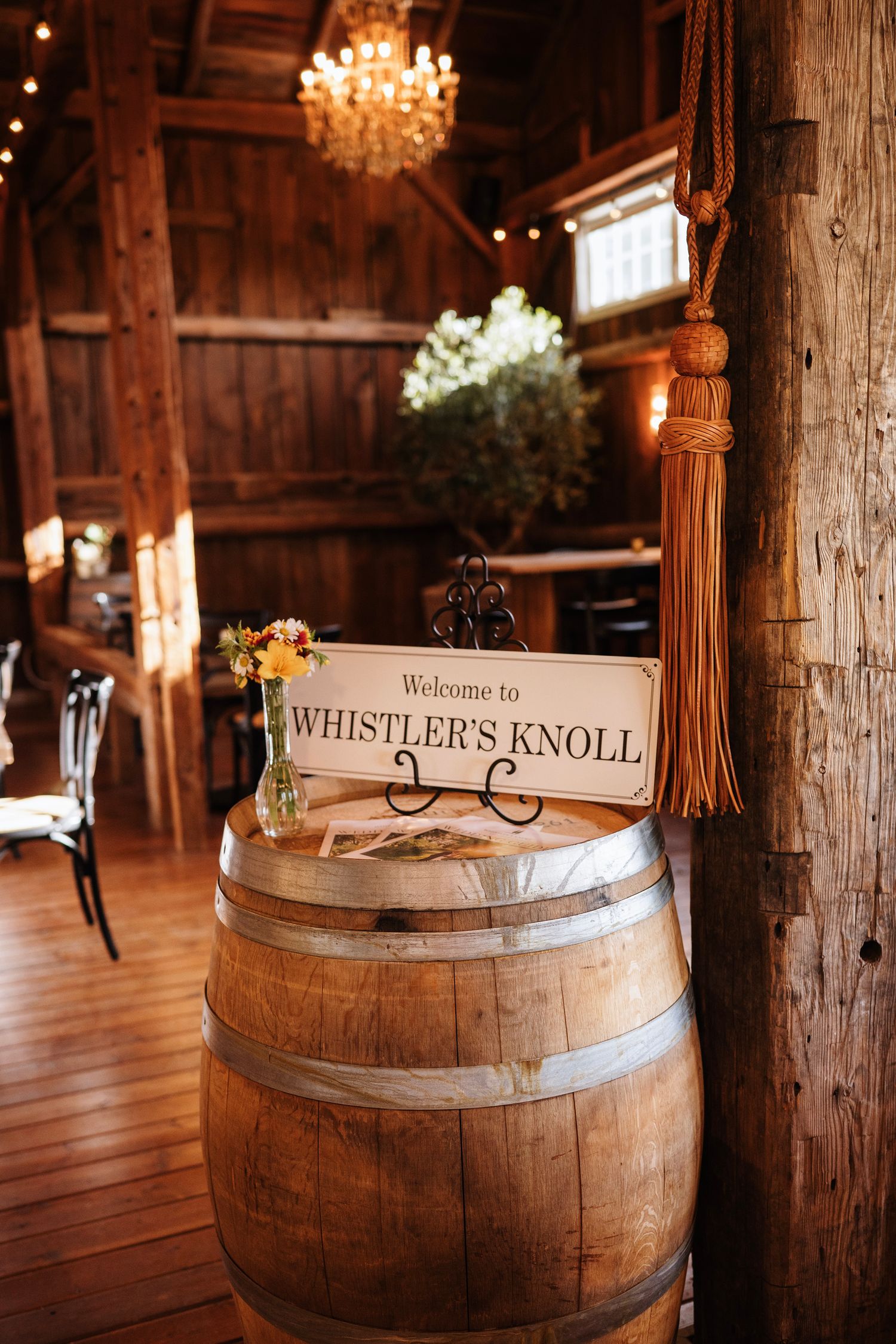 Rustic barn wedding venue with a wooden barrel displaying a Welcome to Whistler's Knoll sign, chandelier, and string lights.
