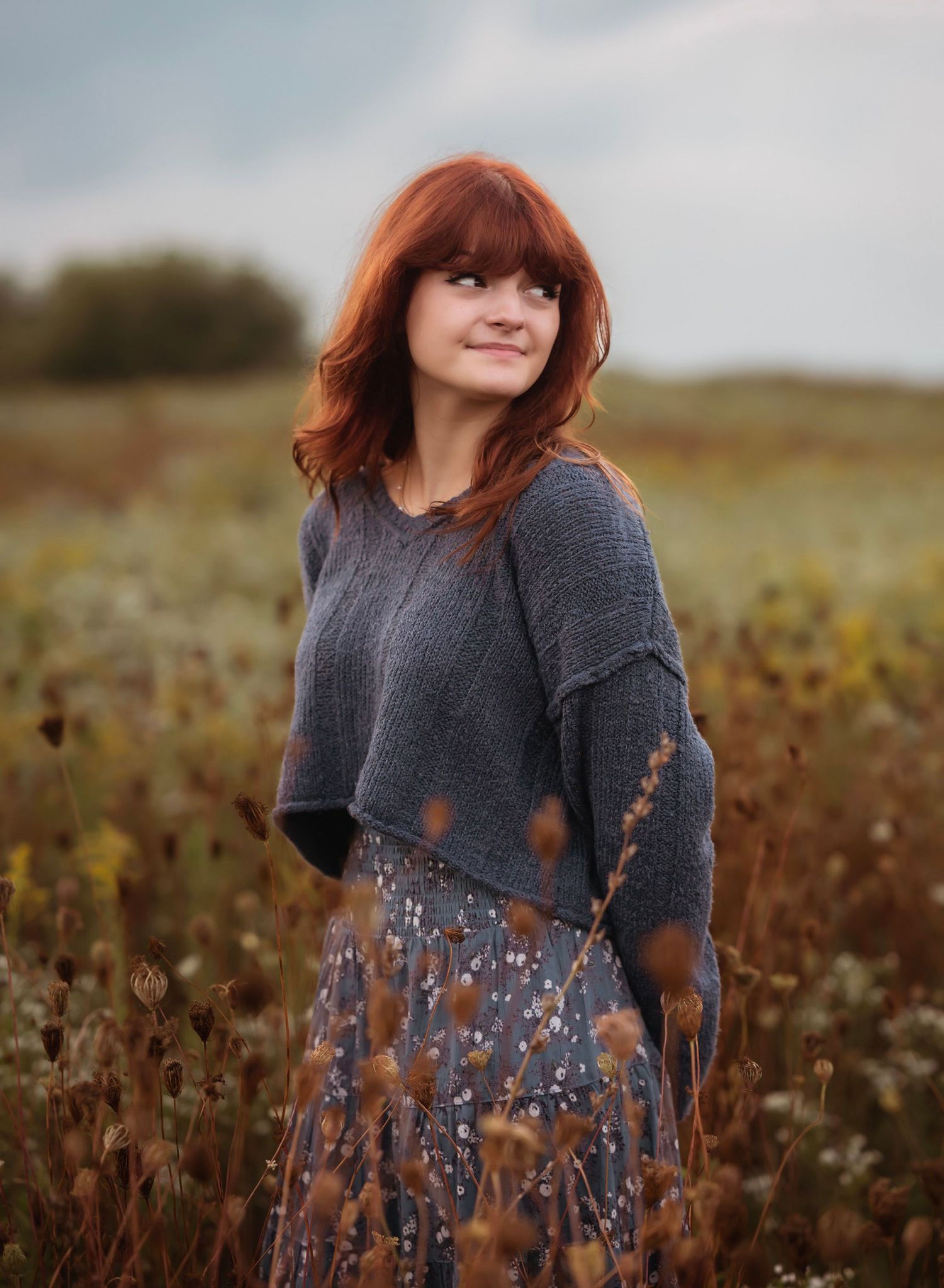 A person in a grey sweater and floral dress stands in a rustic autumn field at sunset.