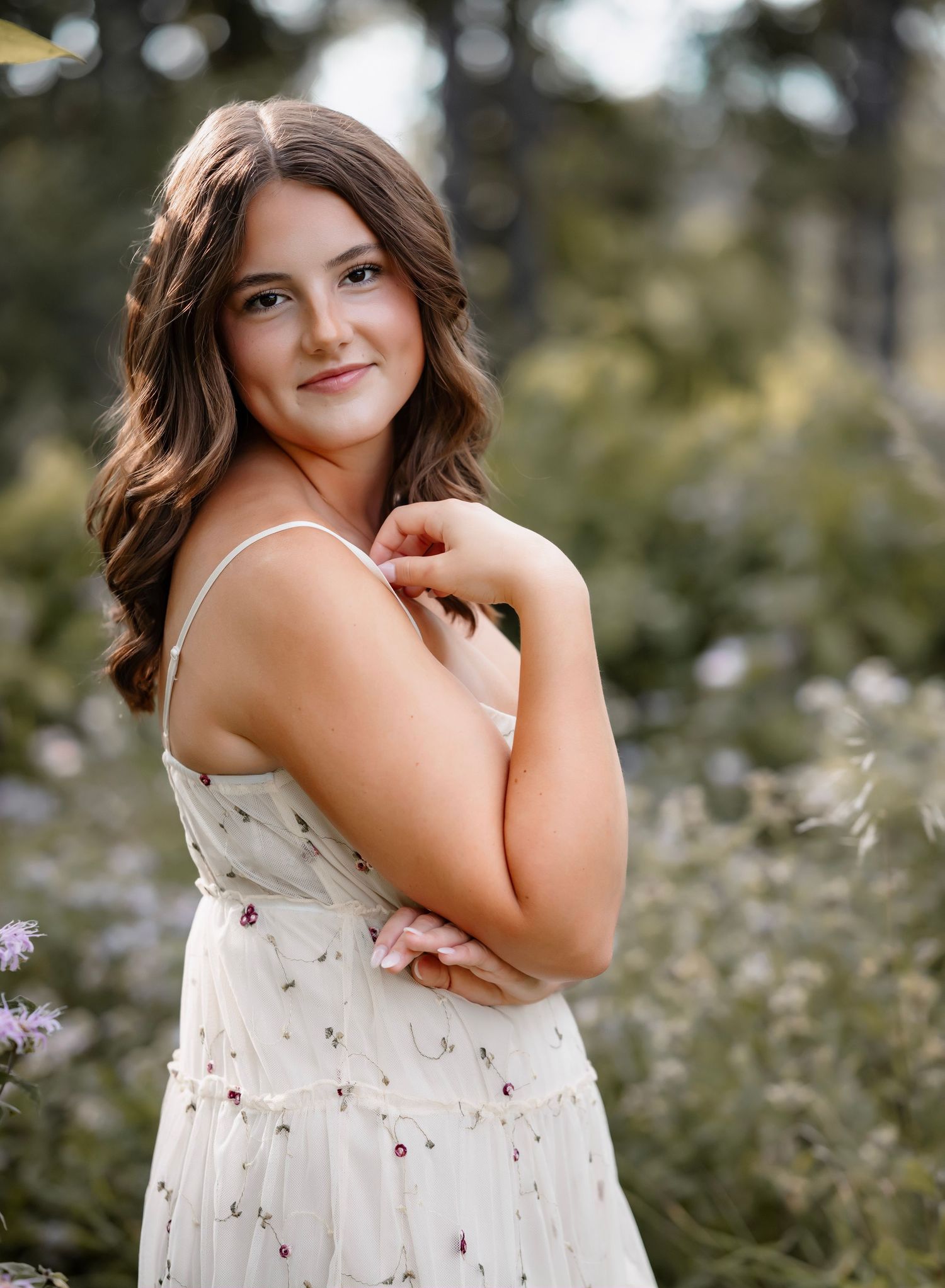 A person in a white floral sundress poses outdoors in a field of purple wildflowers during a summer photoshoot.