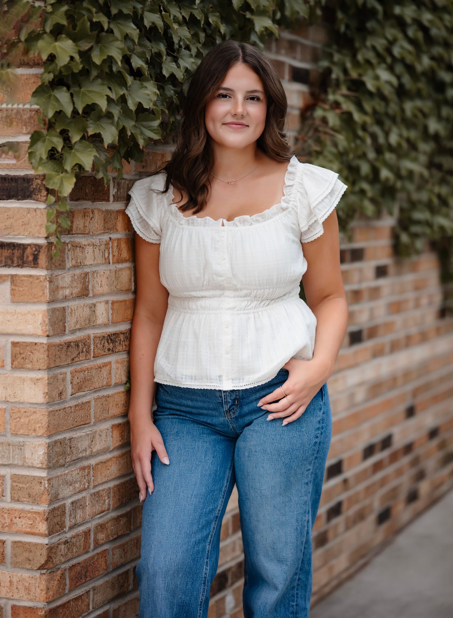 Woman in white peasant top and blue jeans poses against brick wall with ivy backdrop.