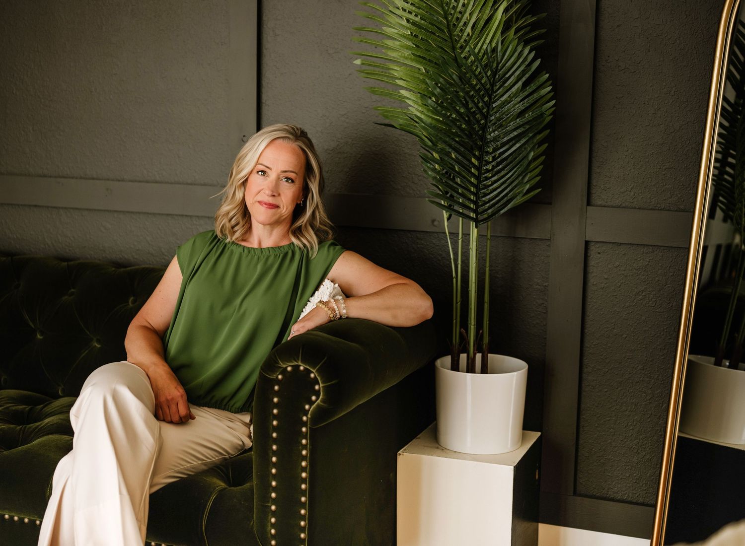 Person in elegant green sleeveless top sits on dark chair near potted palm plant against dark paneled wall.