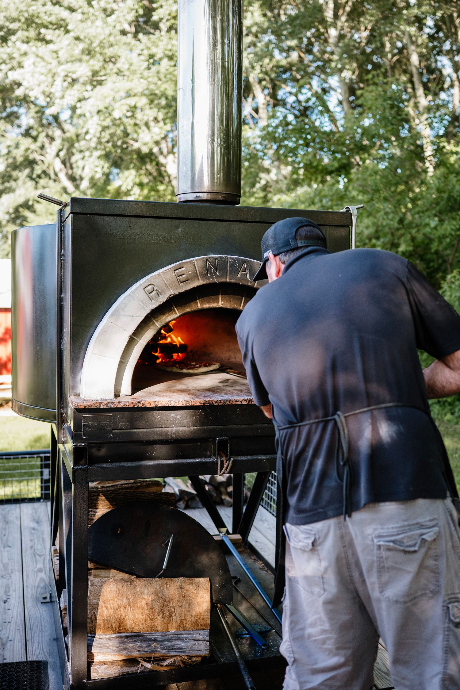 Man tending a wood-fired pizza oven outdoors, with glowing flames visible inside the arched metal oven.