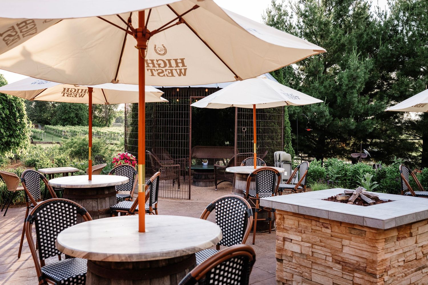 Outdoor patio with High West branded umbrellas, bistro tables, wicker chairs, and a stone fire pit surrounded by lush greenery.