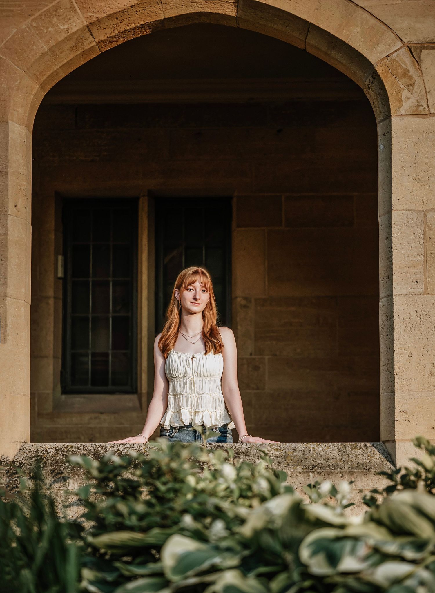 A person in a white lace dress poses thoughtfully under stone archways surrounded by green foliage.