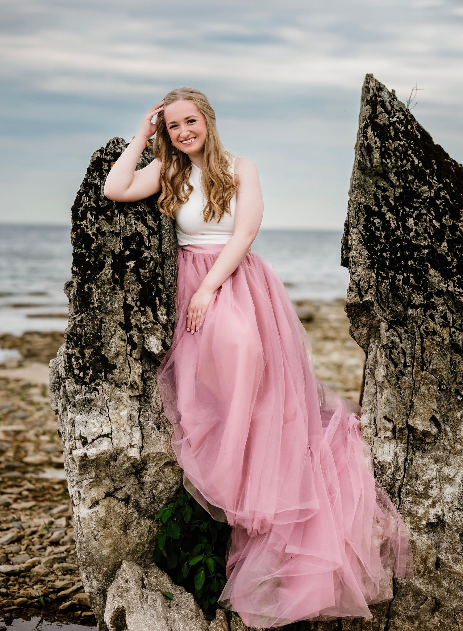 A figure in a flowing pink tulle gown poses gracefully against dramatic coastal rock formations at sunset.