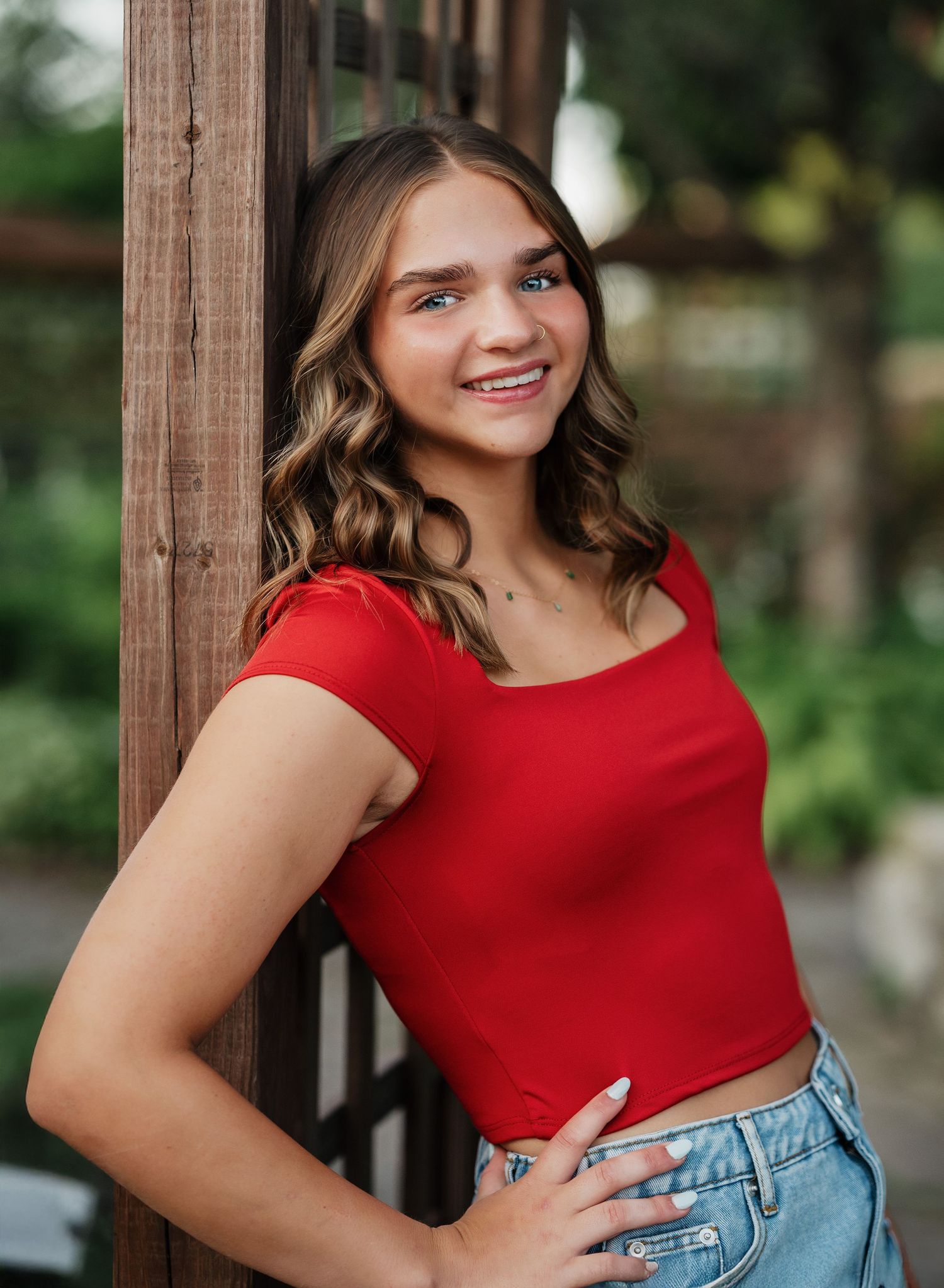 Smiling woman in red top and jeans stands against wooden post outdoors with natural background.