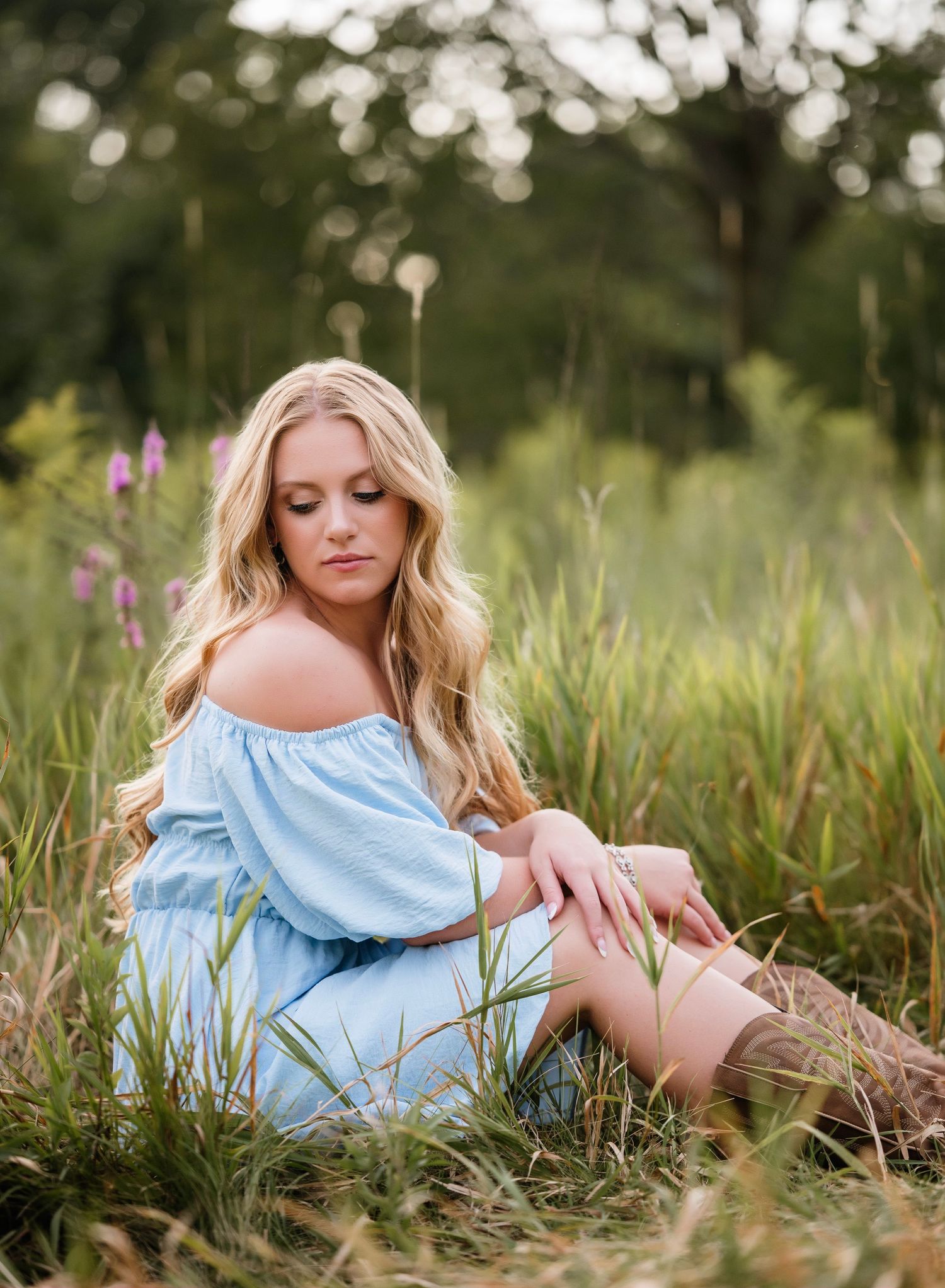 Ethereal portrait in a summer meadow features a light blue off-shoulder dress amid tall grass and wildflowers at golden hour.