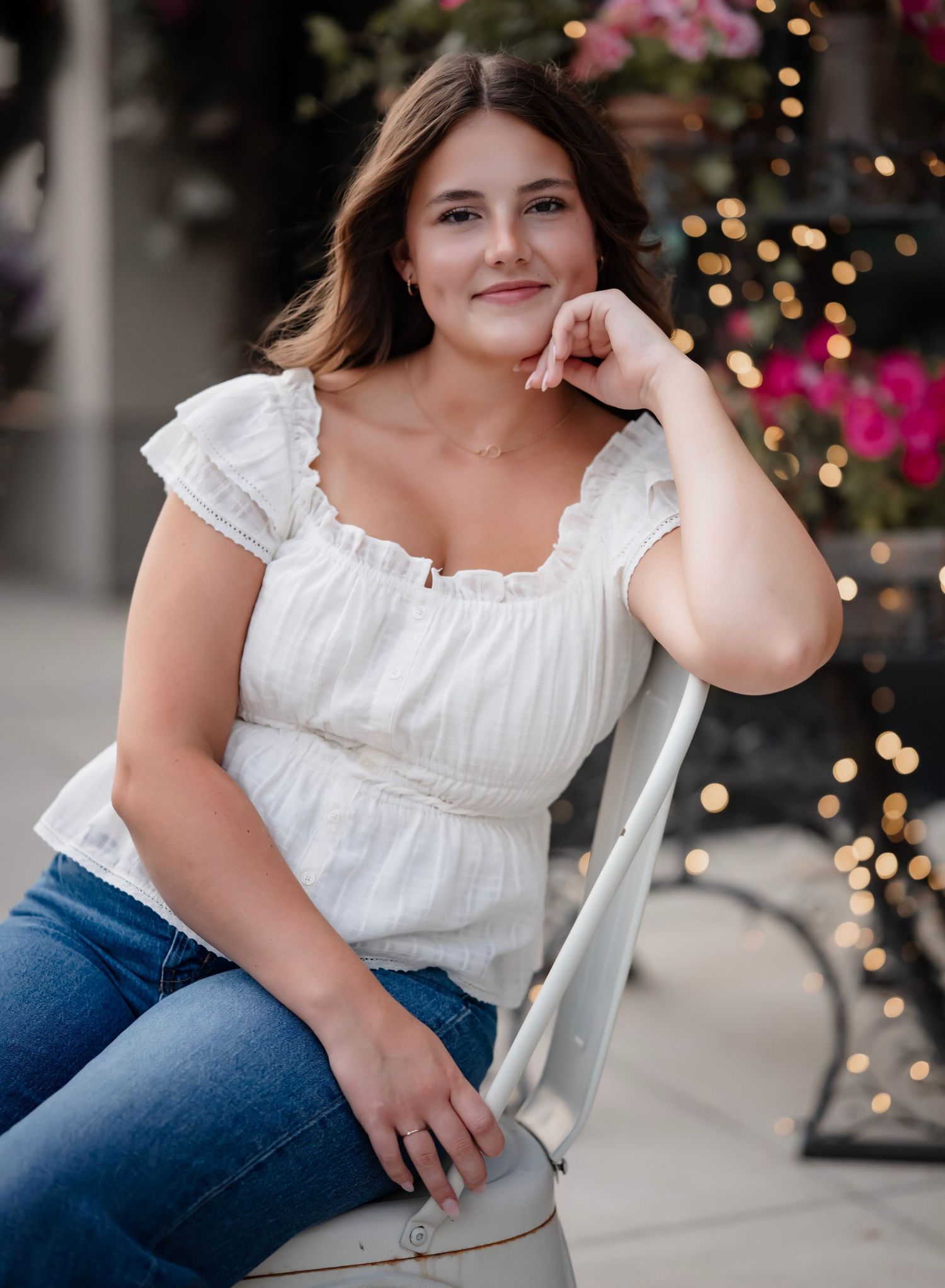A person in a white ruffled top and blue jeans sits casually on a white chair with twinkling lights in the background.