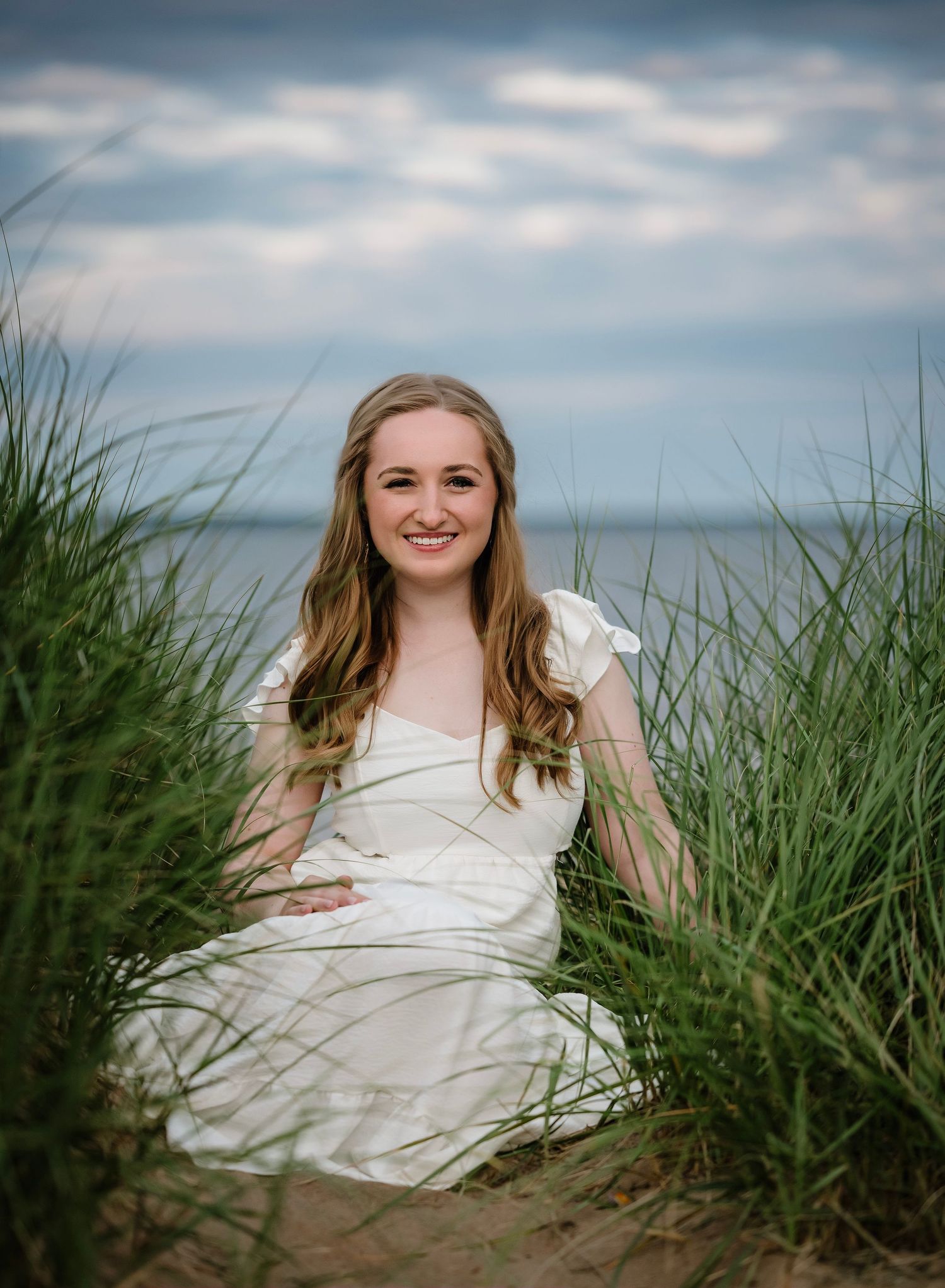 A person in a white dress sits among tall beach grass with a moody cloudy sky and ocean in the background.