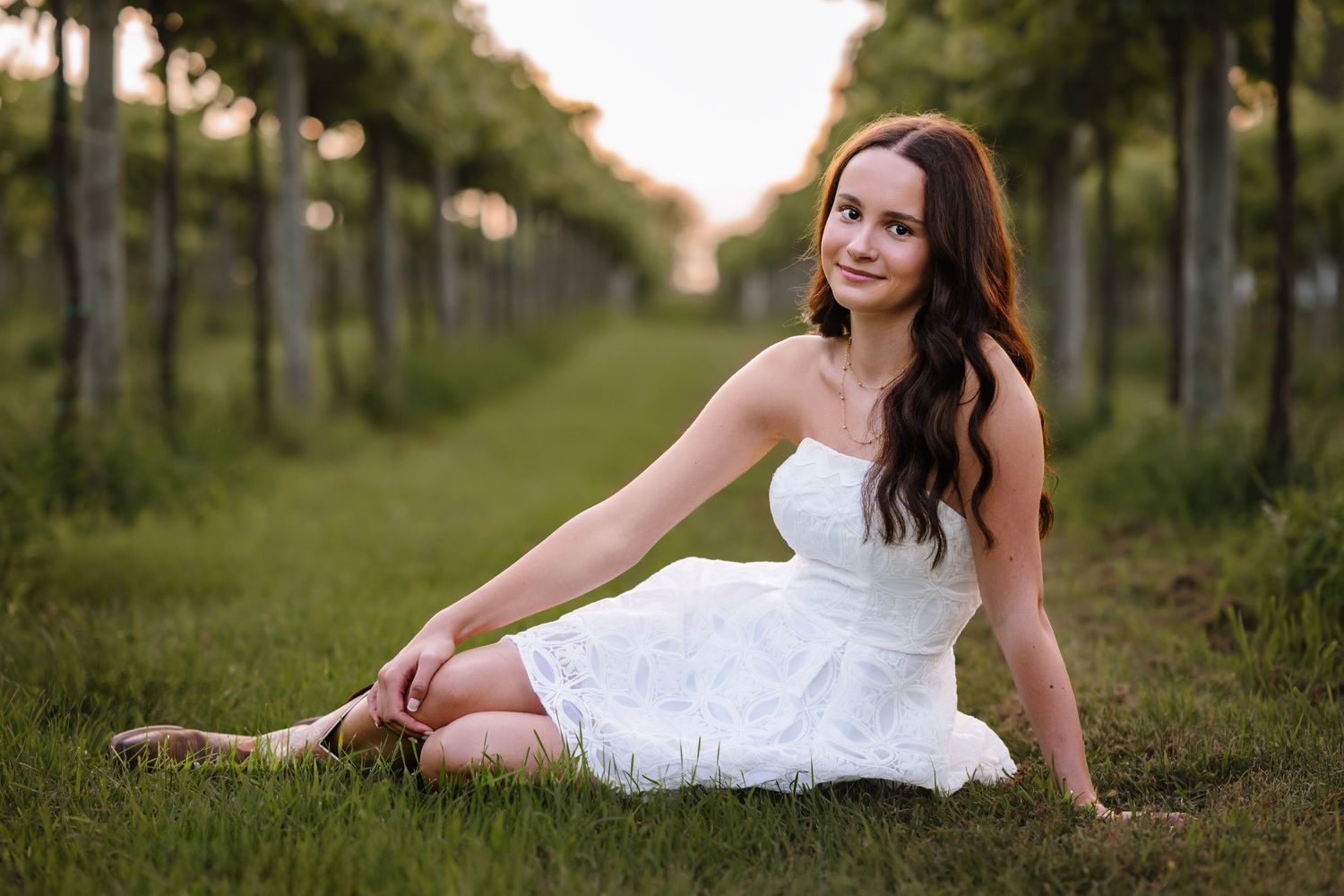 Young woman in white lace dress sitting gracefully on grass between vineyard rows at golden hour.
